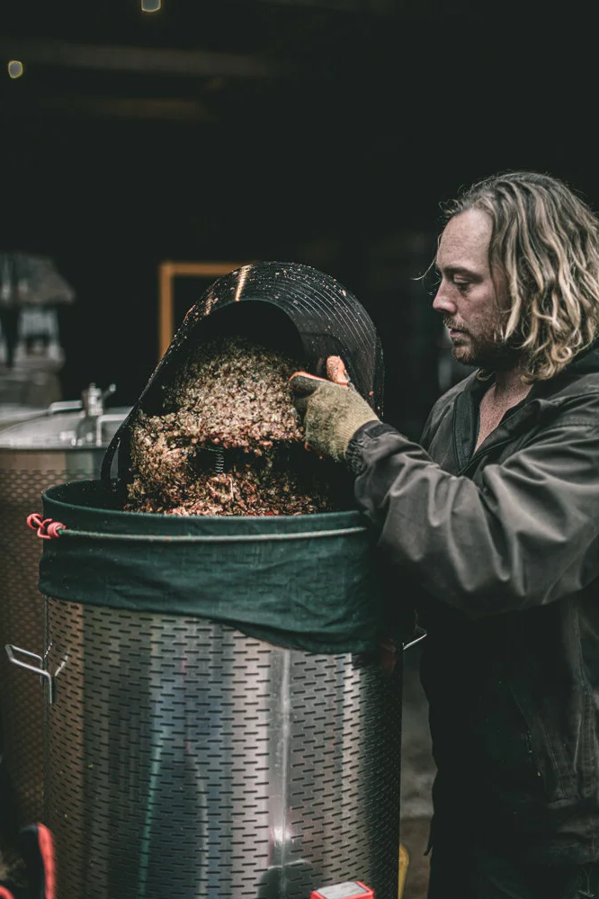Environmental portrait of a drinks producer emptying prepared ingredients into stainless-steel processing equipment, documenting the physical work, machinery and specialist production process behind Sheppys Cider