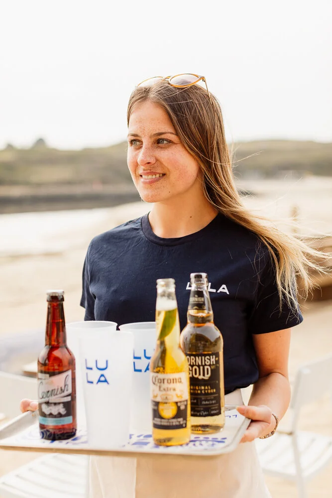 Commercial hospitality portrait of a smiling server carrying a tray of bottled drinks and cups in an outdoor setting, presenting the person naturally within the practical work of service for Lula Shack in Cornwall.