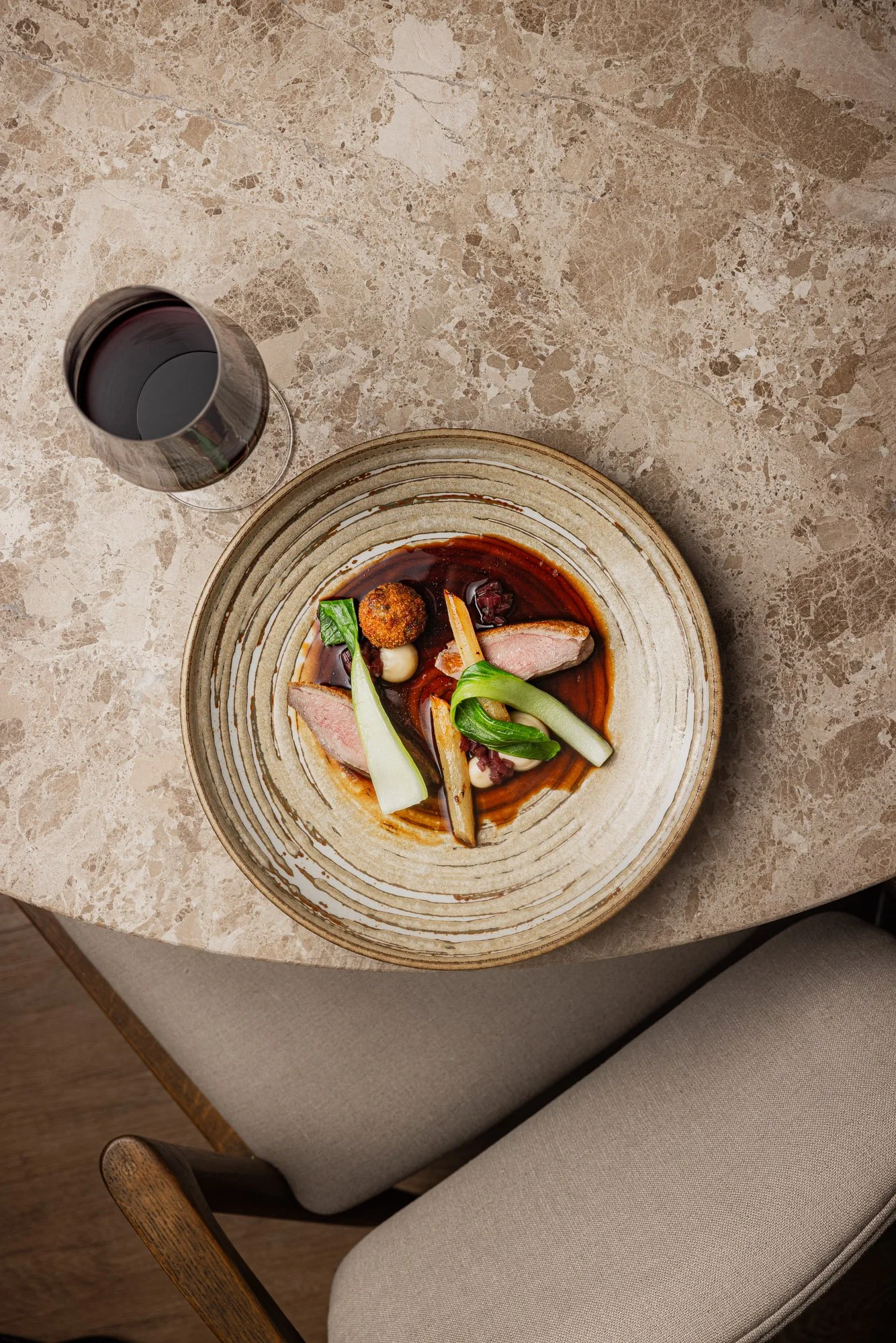 head commercial food photograph of duck breast and roasted vegetable on a ceramic plate beside a wine glass and upholstered dining chair, using geometric composition to place the food within the restaurant setting at the Feather Hotel in Oxfordshire.