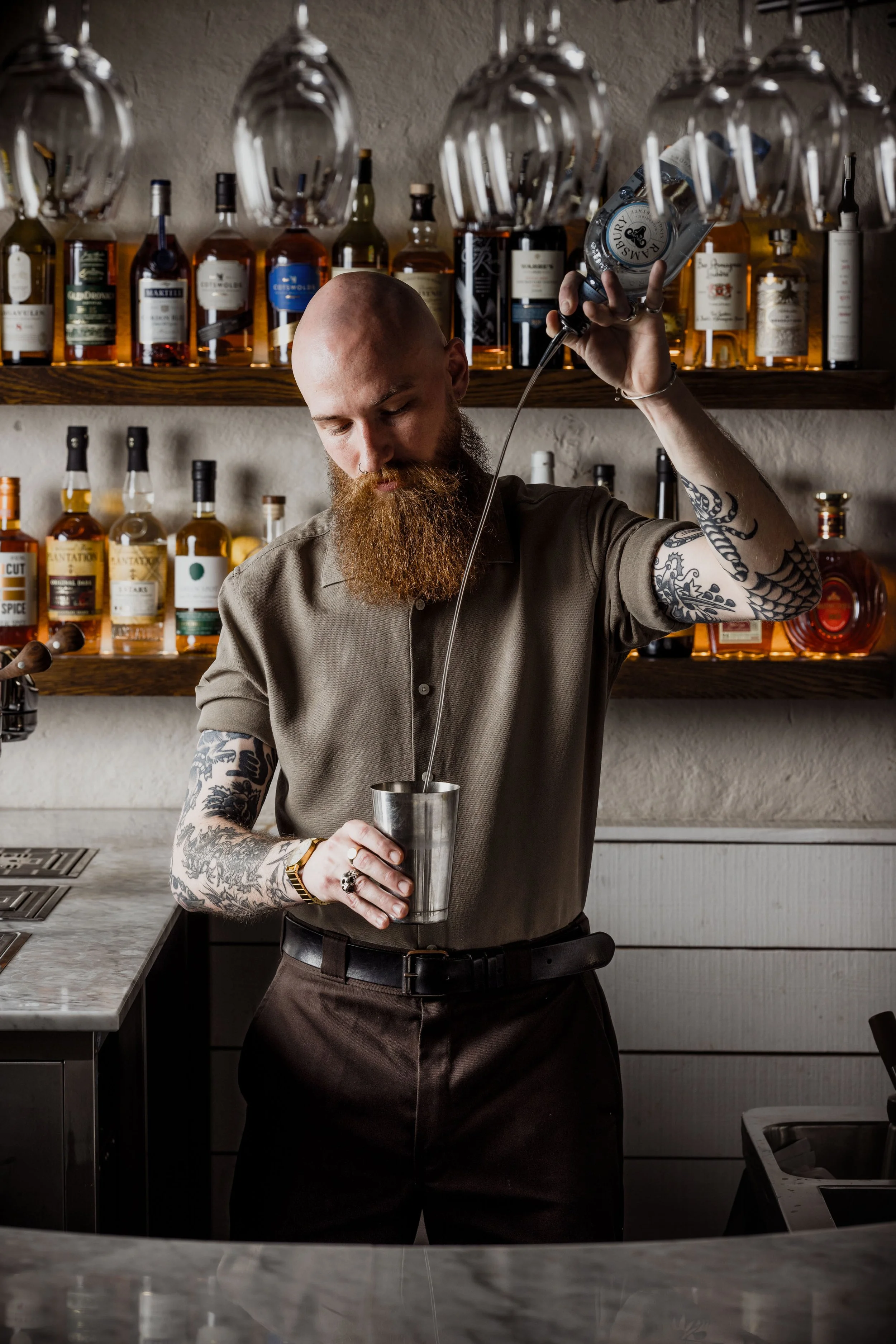 Commercial bartender portrait showing a tattooed bar professional performing a controlled long pour into a metal mixing vessel, framed by shelves of spirits to communicate skill, concentration and the craft of cocktail service.