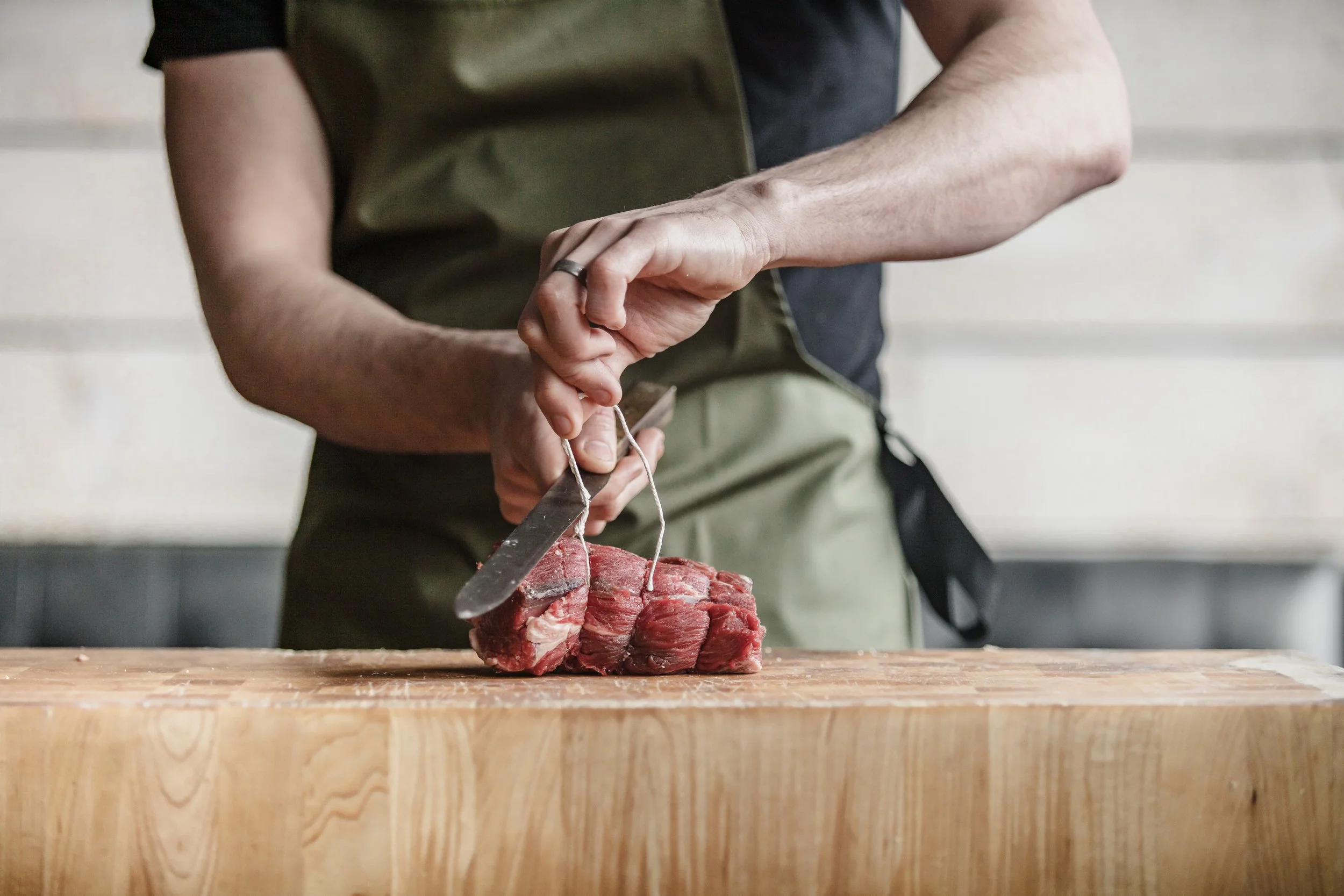 Close commercial photograph of a Pasture Cardiff's butcher slicing a rolled cut of meat on a pale wooden block, focusing on controlled hand movement, knife work and the texture of the prepared meat.