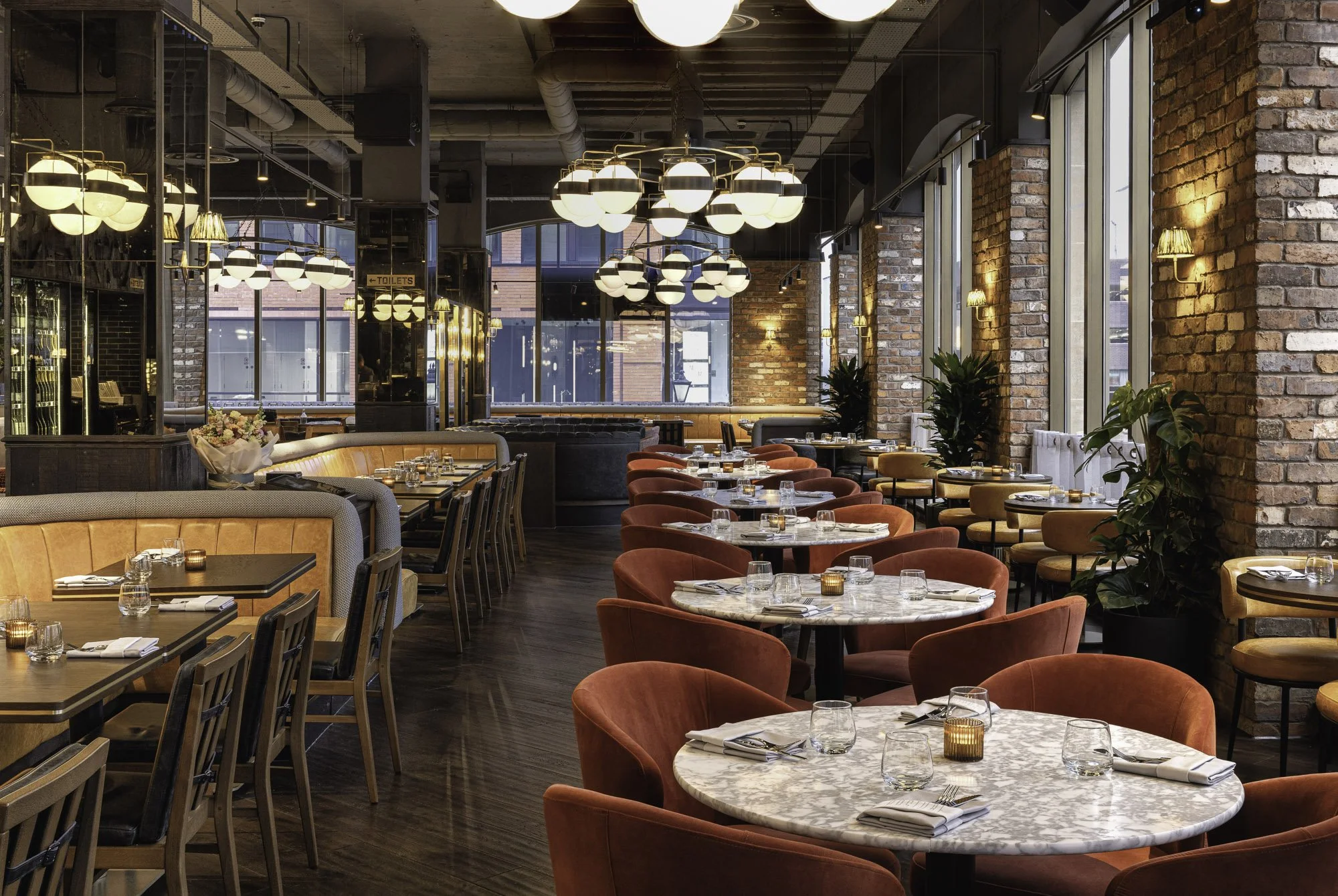 Wide commercial restaurant interior for Pasture Restaurant Group, showing red upholstered chairs, marble tables, exposed brickwork, mirrors and layered ceiling lights.