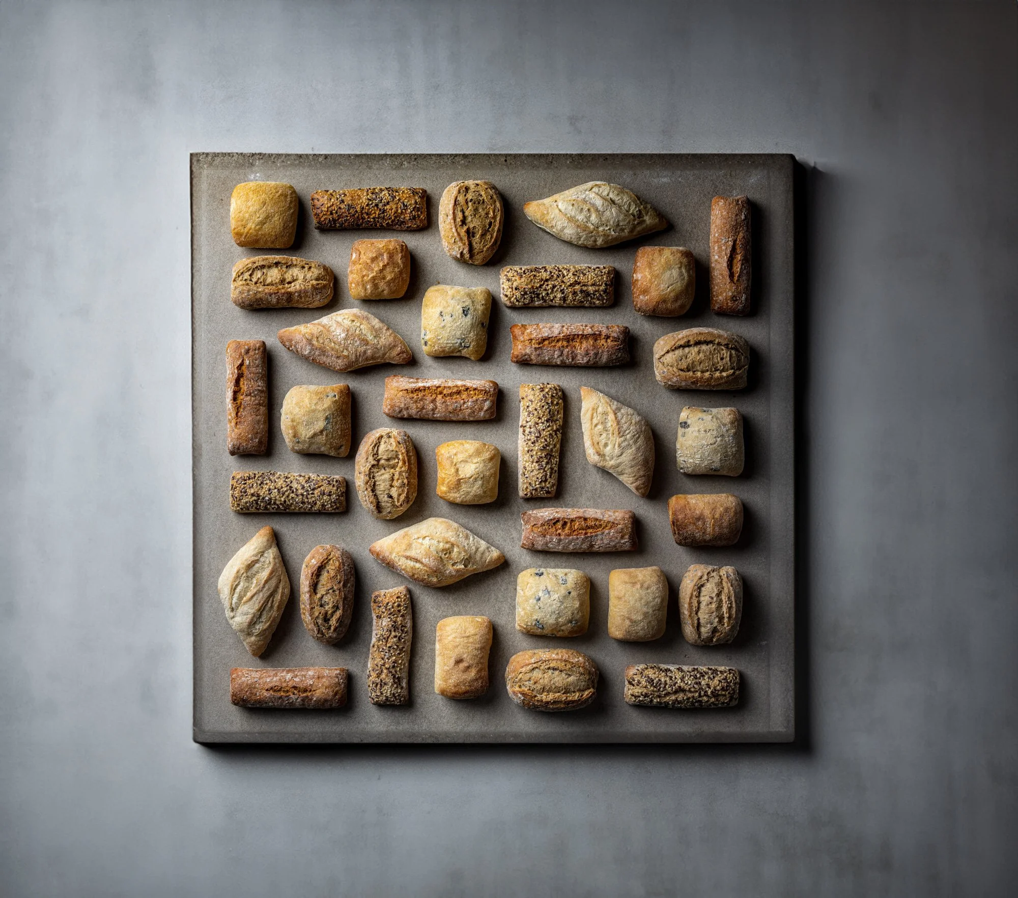 Overhead commercial bakery photograph of an organised range of rolls, loaves and shaped breads arranged across a grey surface, presenting the breadth and consistency of a bakery product collection.