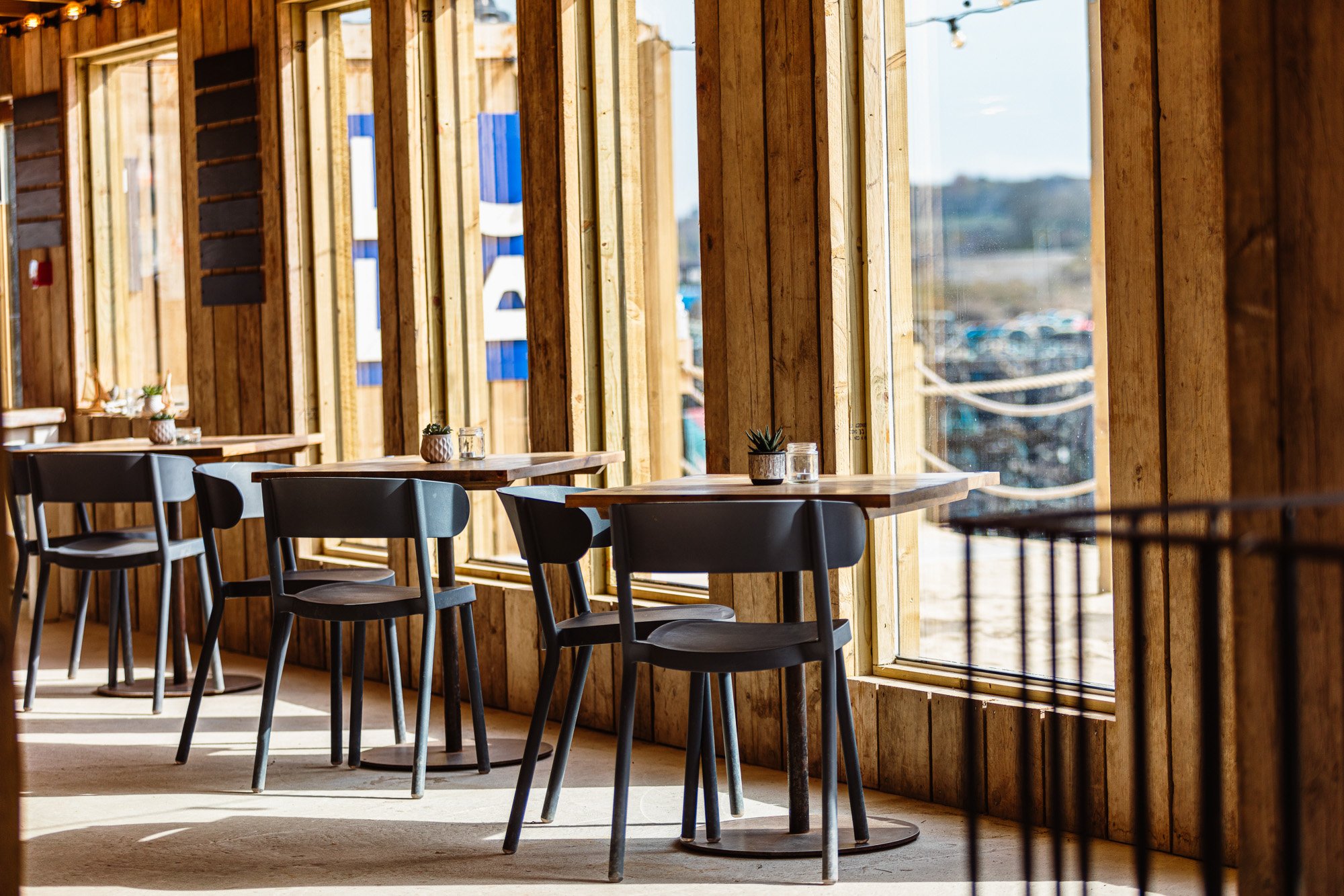 Hospitality interior photograph of a timber-lined restaurant beside tall windows, with black chairs, simple dining tables and strong window shadows, showing how the architecture frames the surrounding view at Lula Shack in Hayle, Cornwall.