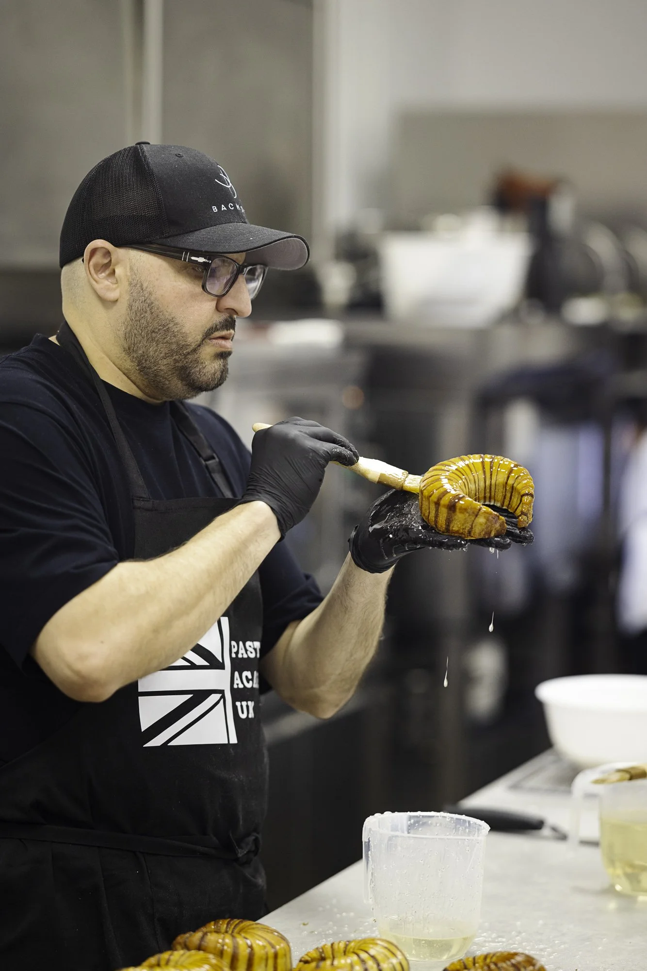 Commercial kitchen photograph of Antonio Bachour using a culinary brush to finish a croissant, focusing on the working hands, protective glove and controlled movement during preparation for Pastry Academy Uk..