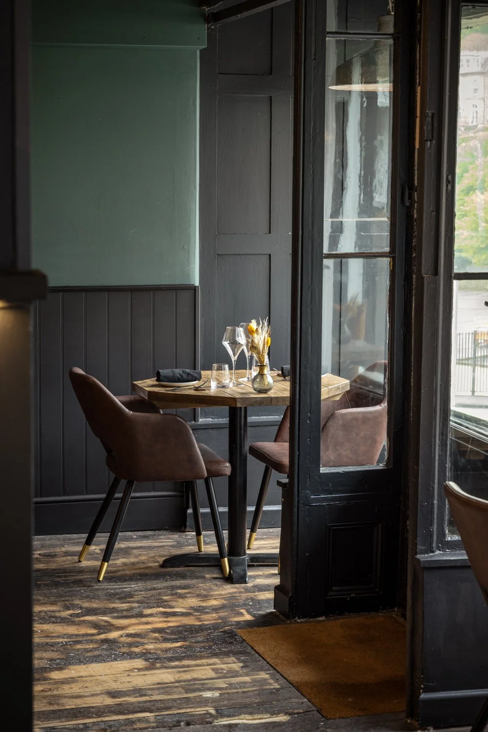 Commercial hospitality interior at Kensington Restaurant in Lynton, Devon, showing a small laid table beside tall windows, a dark doorway and restrained architectural details.