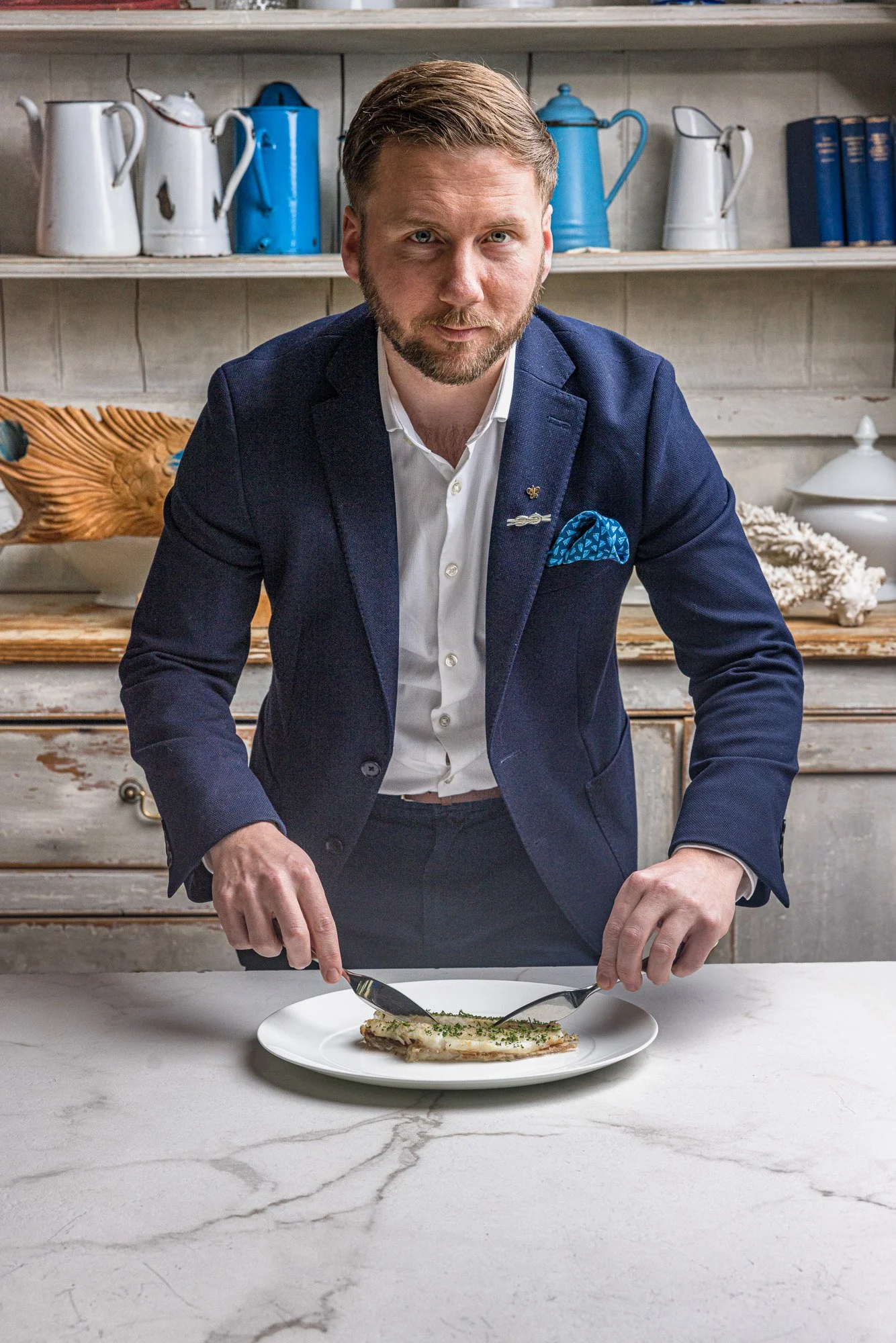 Commercial portrait of a smartly dressed restaurant professional finishing a plated dish at the table, combining a formal business appearance with direct involvement in the food for Idle Rock in Cornwall.