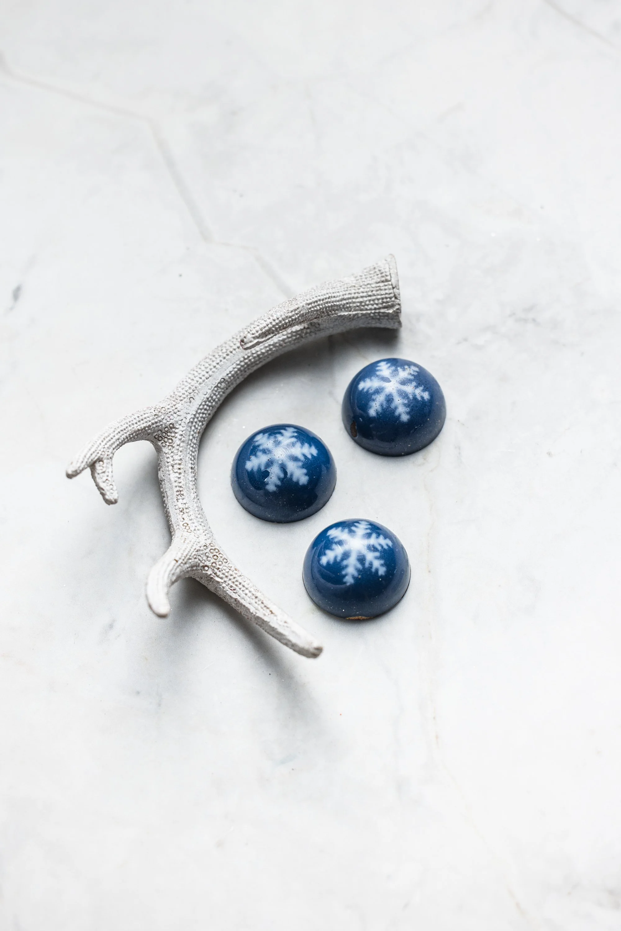 Minimal commercial chocolate photograph of glossy blue bonbons placed on a white sculptural prop against a pale marble surface, using negative space and tonal contrast to emphasise colour and finish