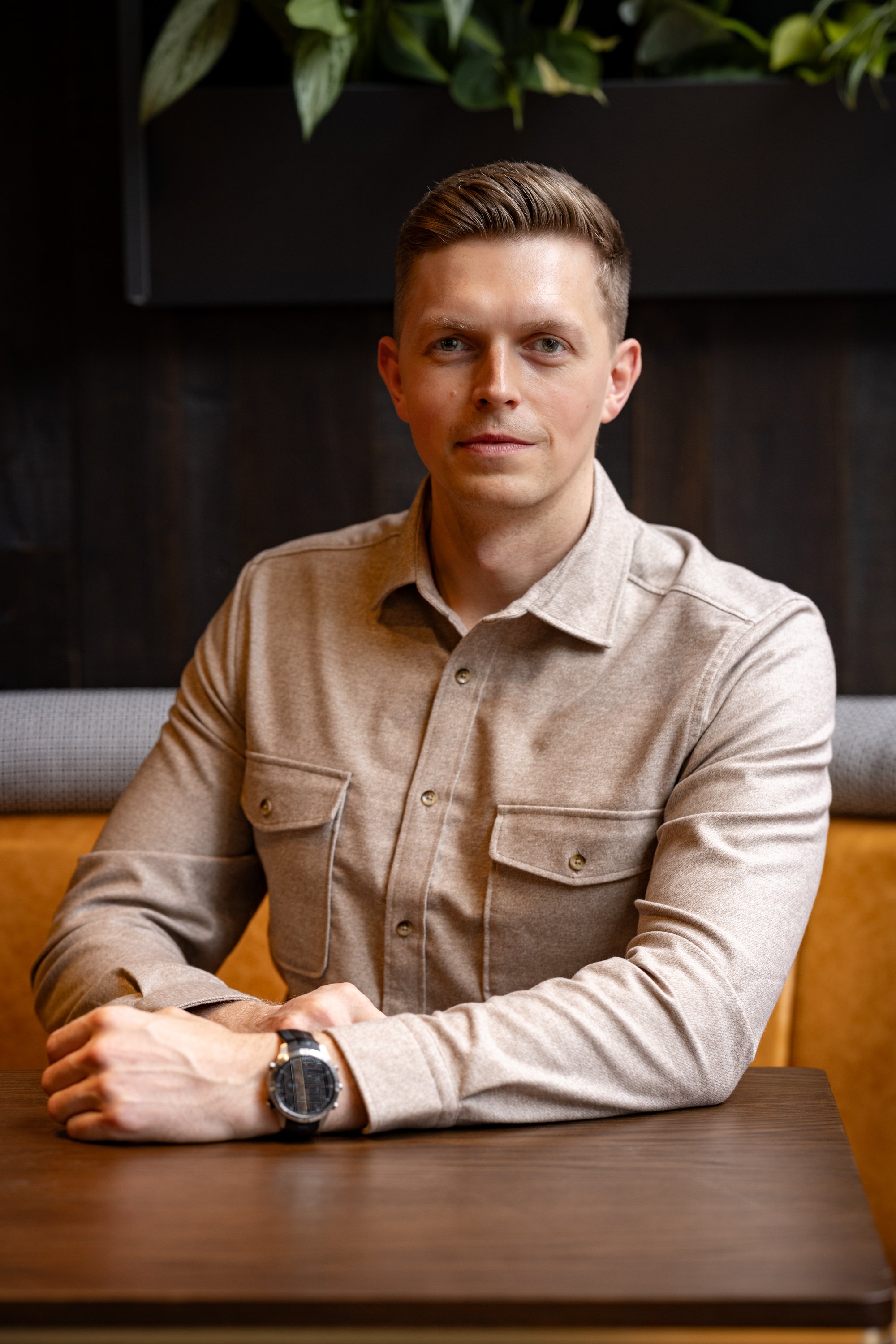 Contemporary corporate portrait of a young businessman seated at a dark table in a warm interior, using a direct expression, relaxed posture and restrained lighting for Pasture Cardiff.
