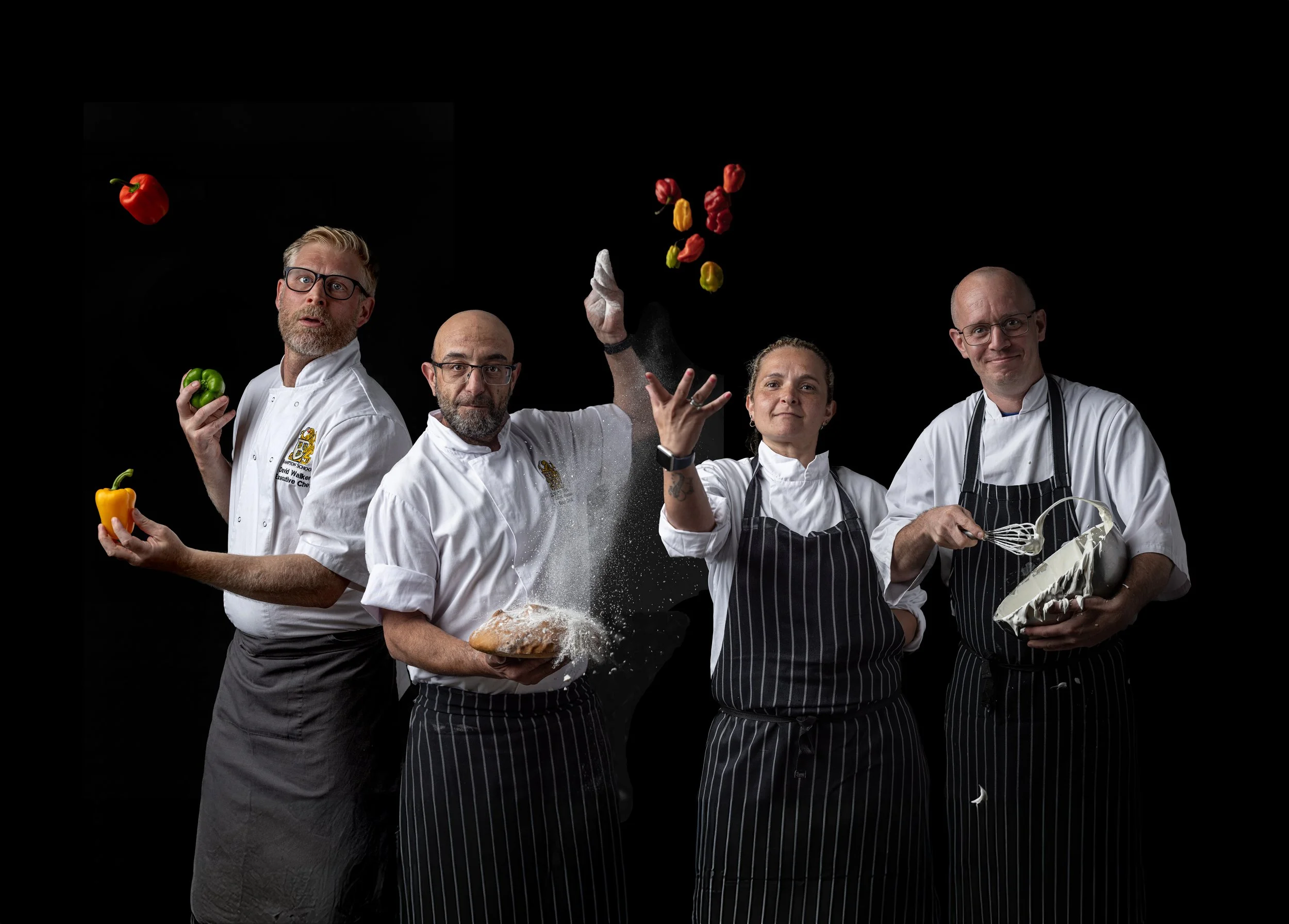 Creative group portrait of four chefs against a black background with colourful ingredients suspended above them, using controlled studio lighting and playful movement to show the personalities behind Hampton School rebranding campaign in London.
