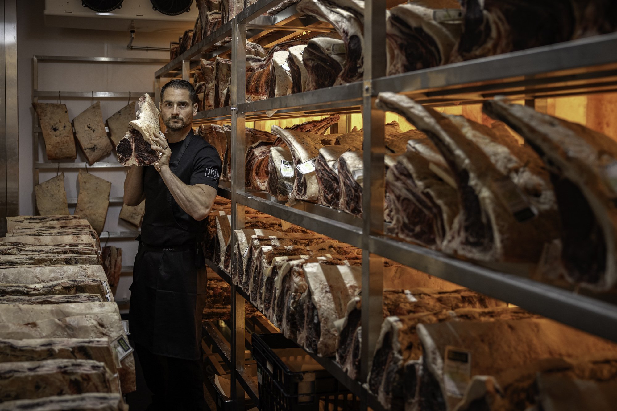 Environmental butchery photograph of a producer standing inside a warmly lit dry-ageing room lined with hanging beef, showing the scale, process and specialist production setting for Pasture in Bristol.