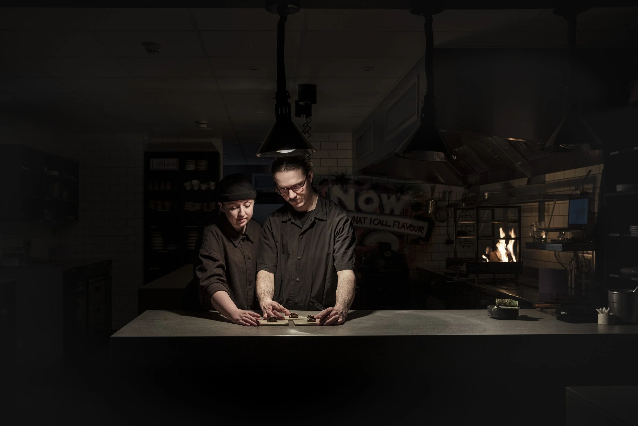 Atmospheric commercial photograph of two chefs working together at a preparation counter in a dark kitchen, using focused overhead lighting to isolate their hands, concentration and collaboration within the wider restaurant space.