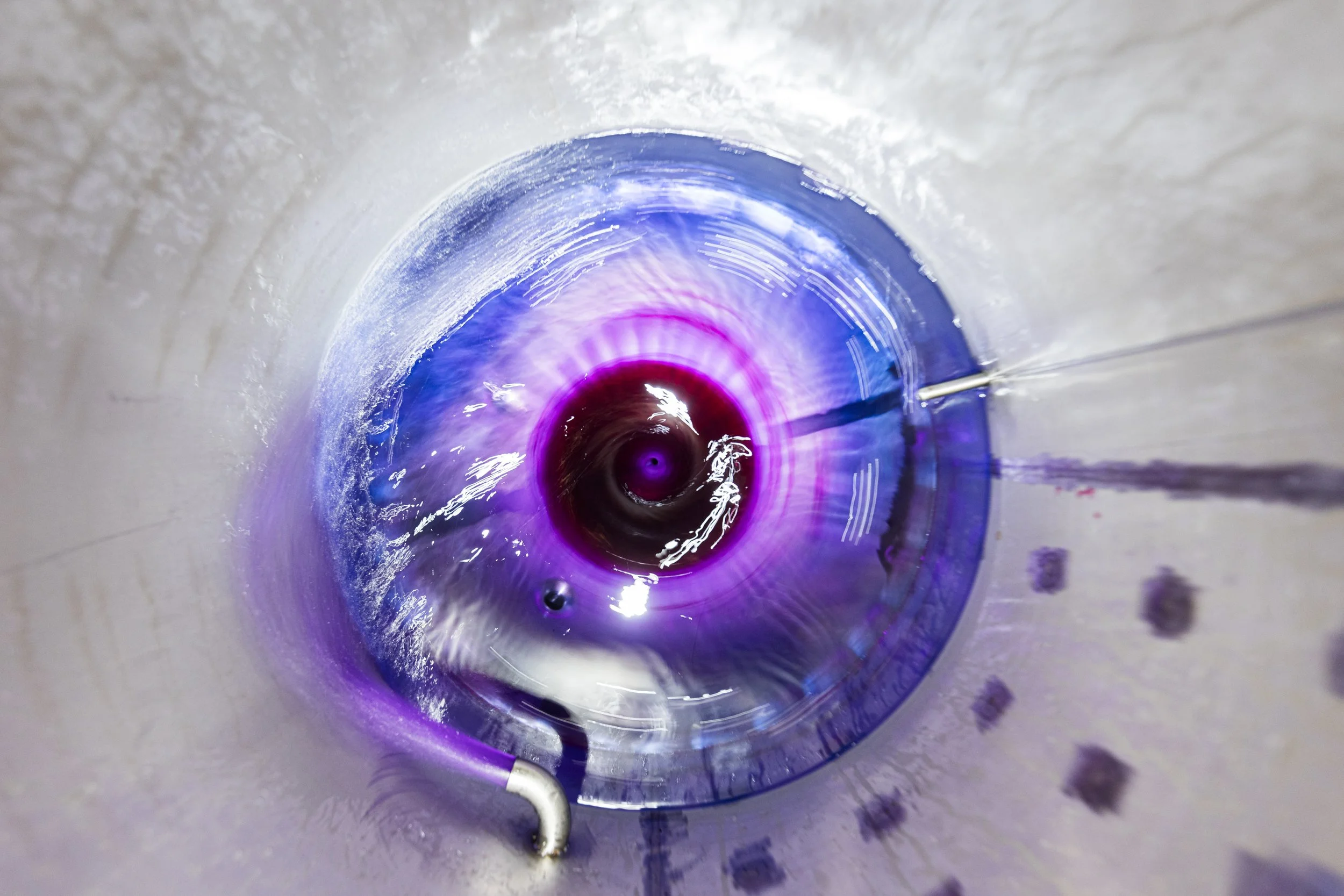Overhead production photograph looking into a stainless-steel tank as vivid purple liquid swirls around a central mixer, using circular movement and reflections to show an active beverage manufacturing process.