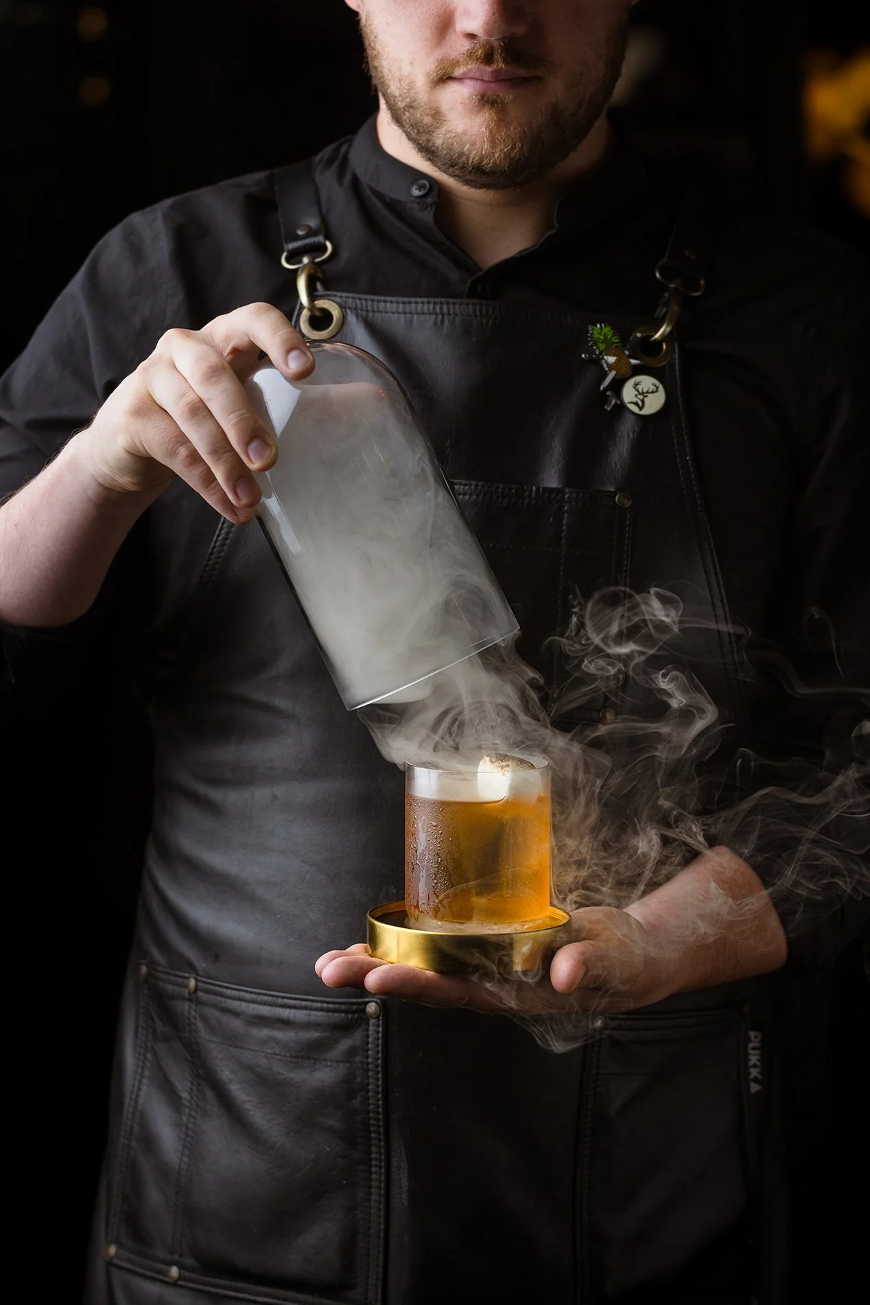 Hospitality drinks photograph of a bartender presenting an amber cocktail surrounded by rising smoke, capturing the theatrical preparation, working hands and controlled bar lighting for Bistro Pierre in Torquay.