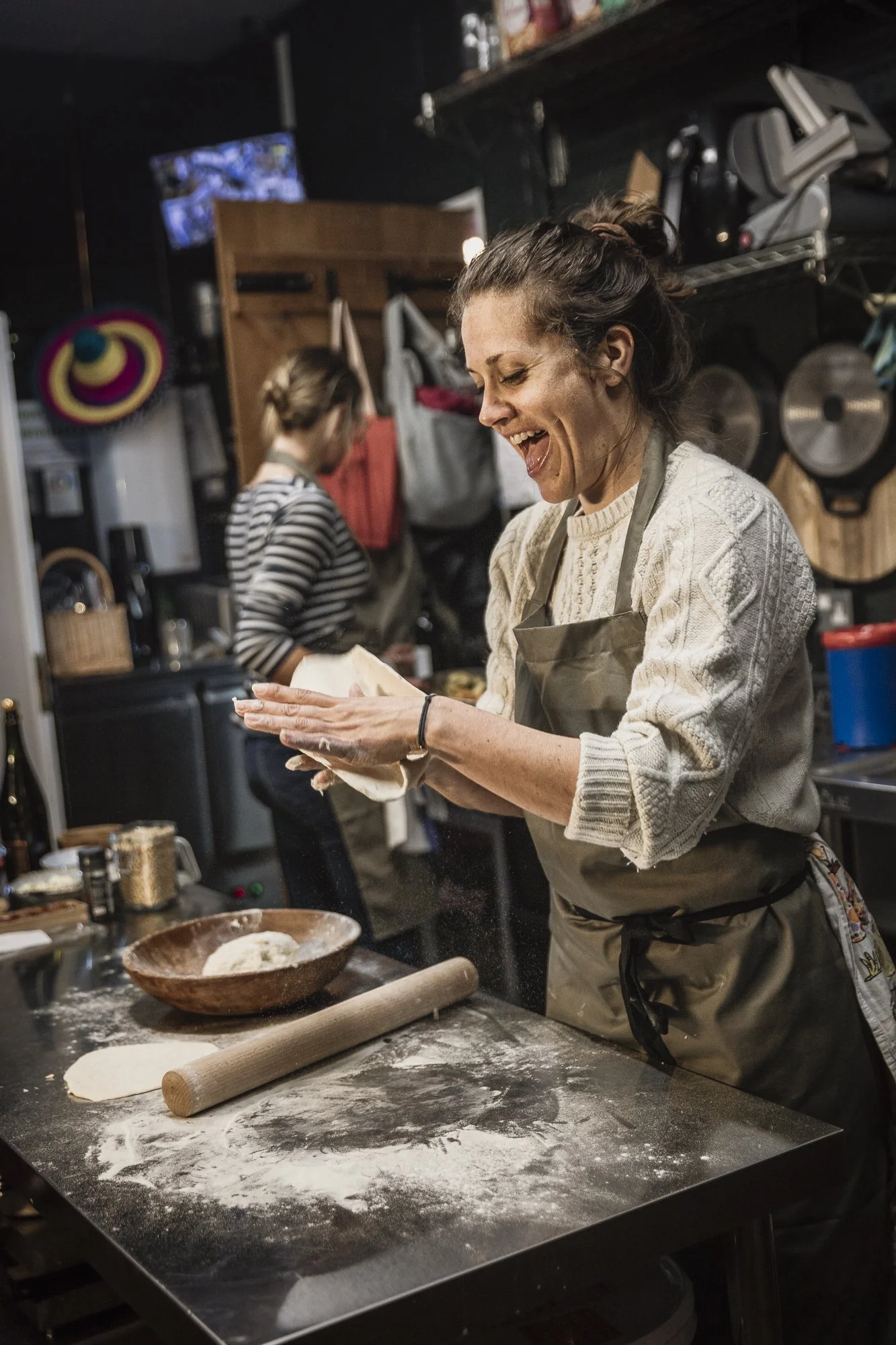 Candid commercial portrait of a woman smiling while working with flour in a busy kitchen, preserving the movement, practical surroundings and unguarded expression that connect her naturally with the work.