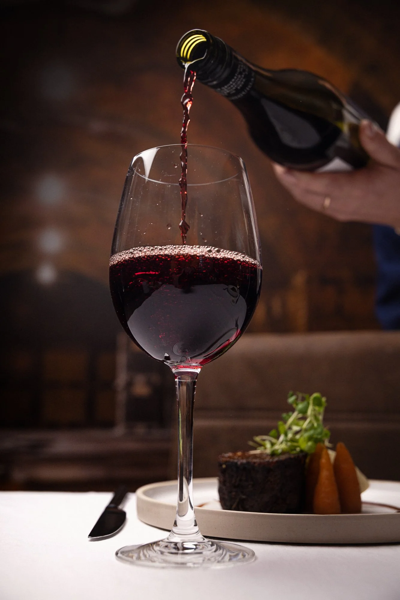Restaurant drinks photograph of red wine being poured into a stemmed glass beside a plated dish, placing the wine within a warm dining setting and capturing the colour and movement of table service.