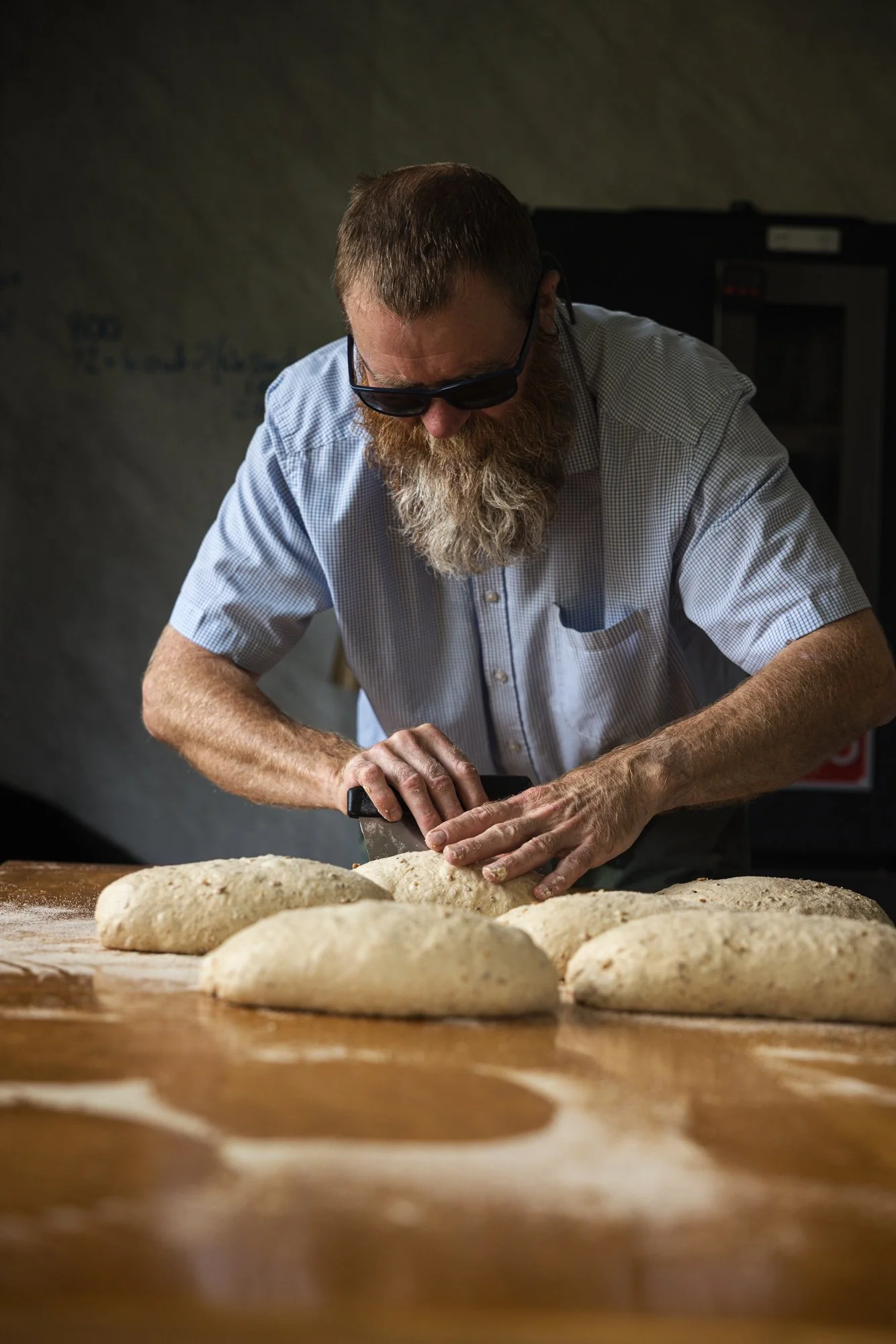 Environmental portrait of an artisan baker leaning over several loaves while kneading dough by hand at a flour-covered bench, using warm directional light to show concentration, technique and the physical character of bread making.