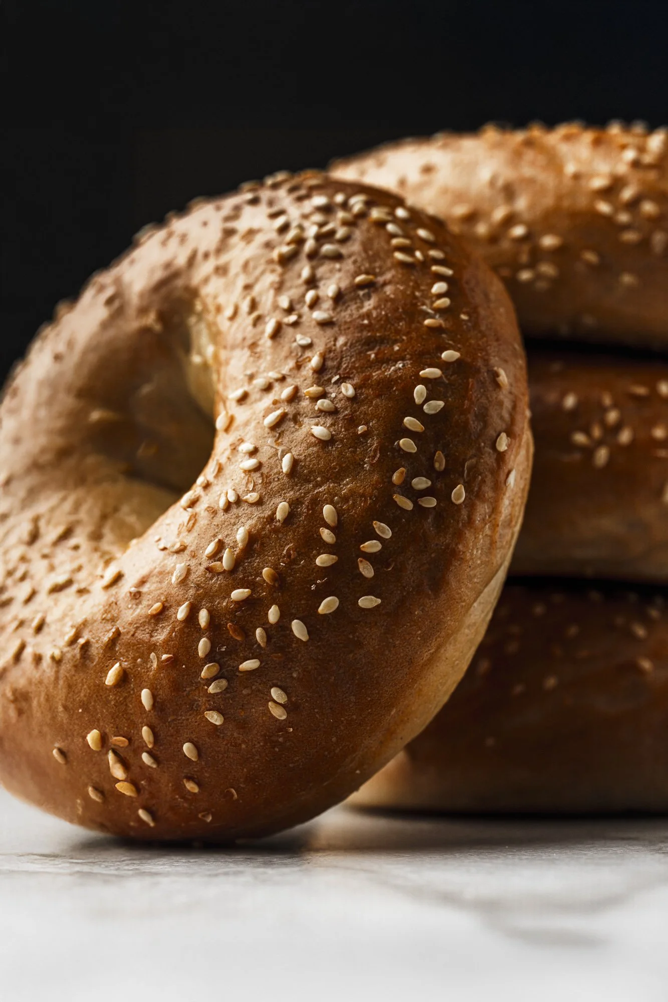 Close commercial food photograph of a sesame-coated bagel with a second bagel softly visible behind it, using low directional lighting to highlight the seeds, golden crust and rounded form.