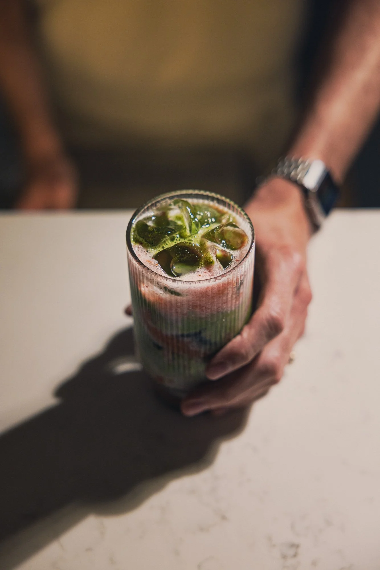 Commercial lifestyle photograph of a hand presenting a textured glass filled with a green iced matcha tea, using hard sunlight and a pale surface to emphasise the colour, ice and fresh garnish for Zuma.