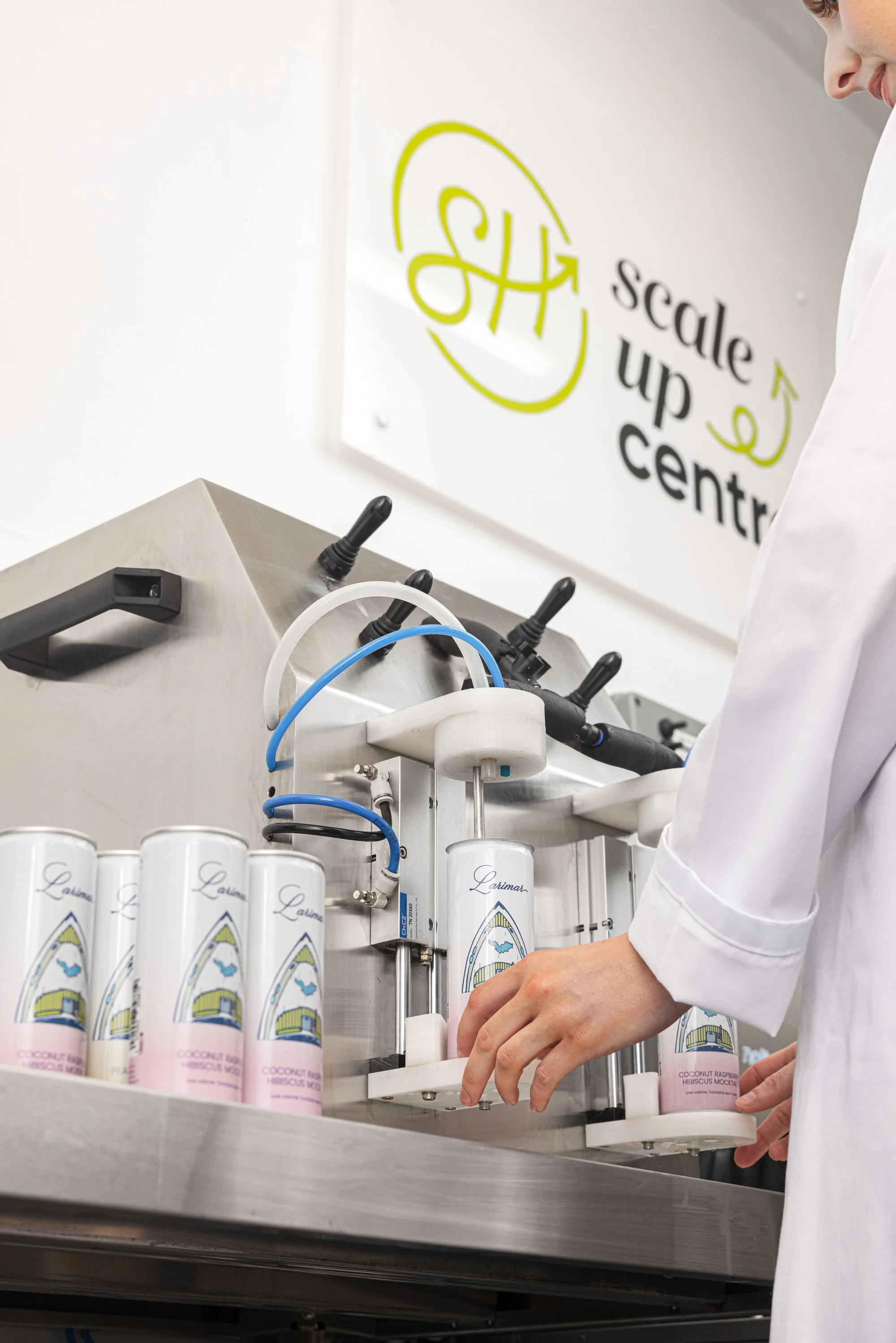 Commercial production photograph of an operator working beside a small drinks canning line, showing labelled cans, filling equipment and the controlled packaging process for soft drink beverage producer.