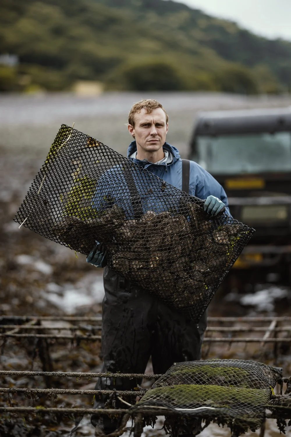 Environmental portrait of an oyster farmer seated among working baskets on a misty shoreline while inspecting a freshly harvested oyster, showing the person, landscape and practical aquaculture work behind Porlock Bay Oyster in Somerset.