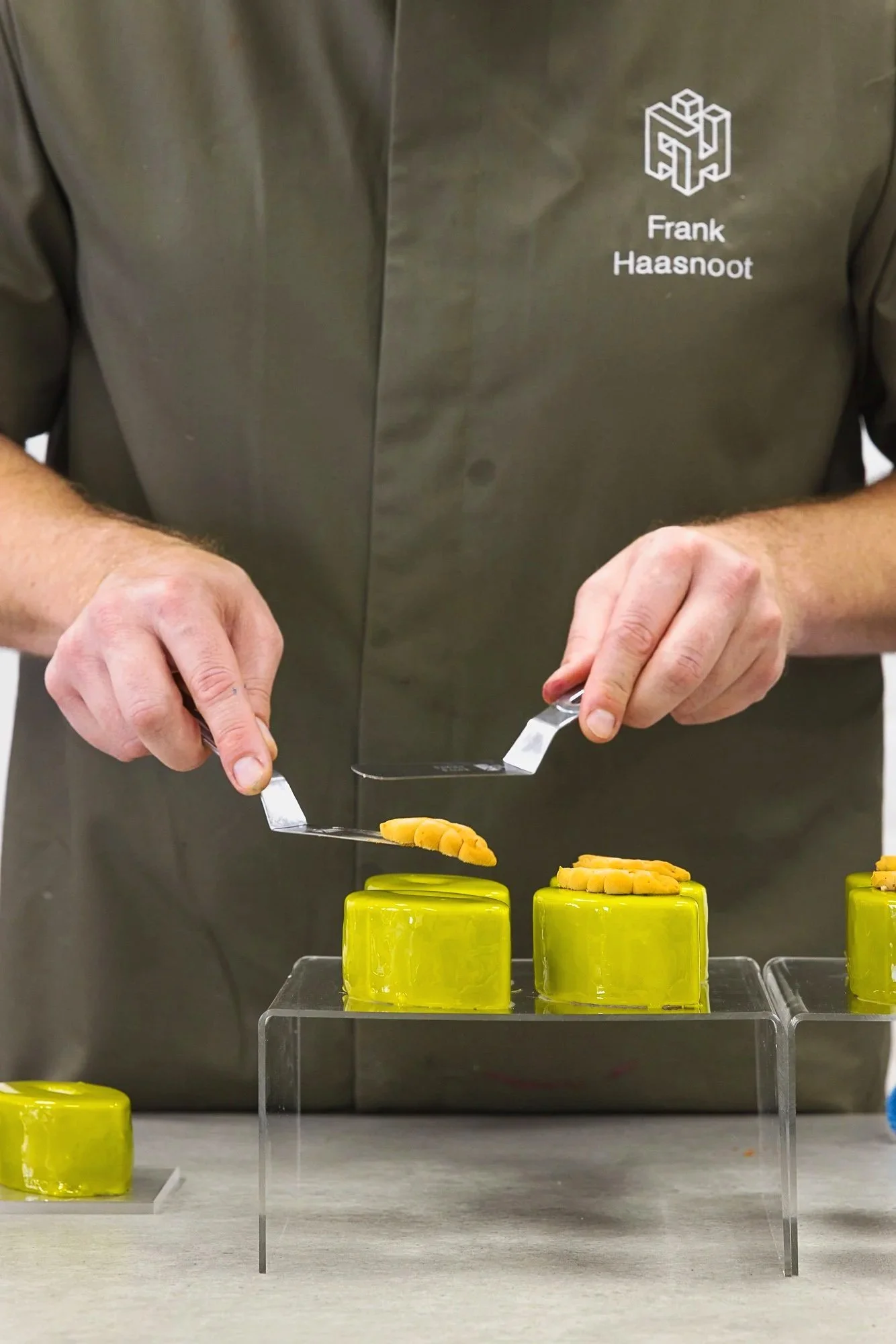 Commercial pastry process photograph showing a Frank Haasnoot cutting neat green individual cakes on clear presentation stands, capturing careful finishing work, symmetry and the precision of professional pâtisserie production.