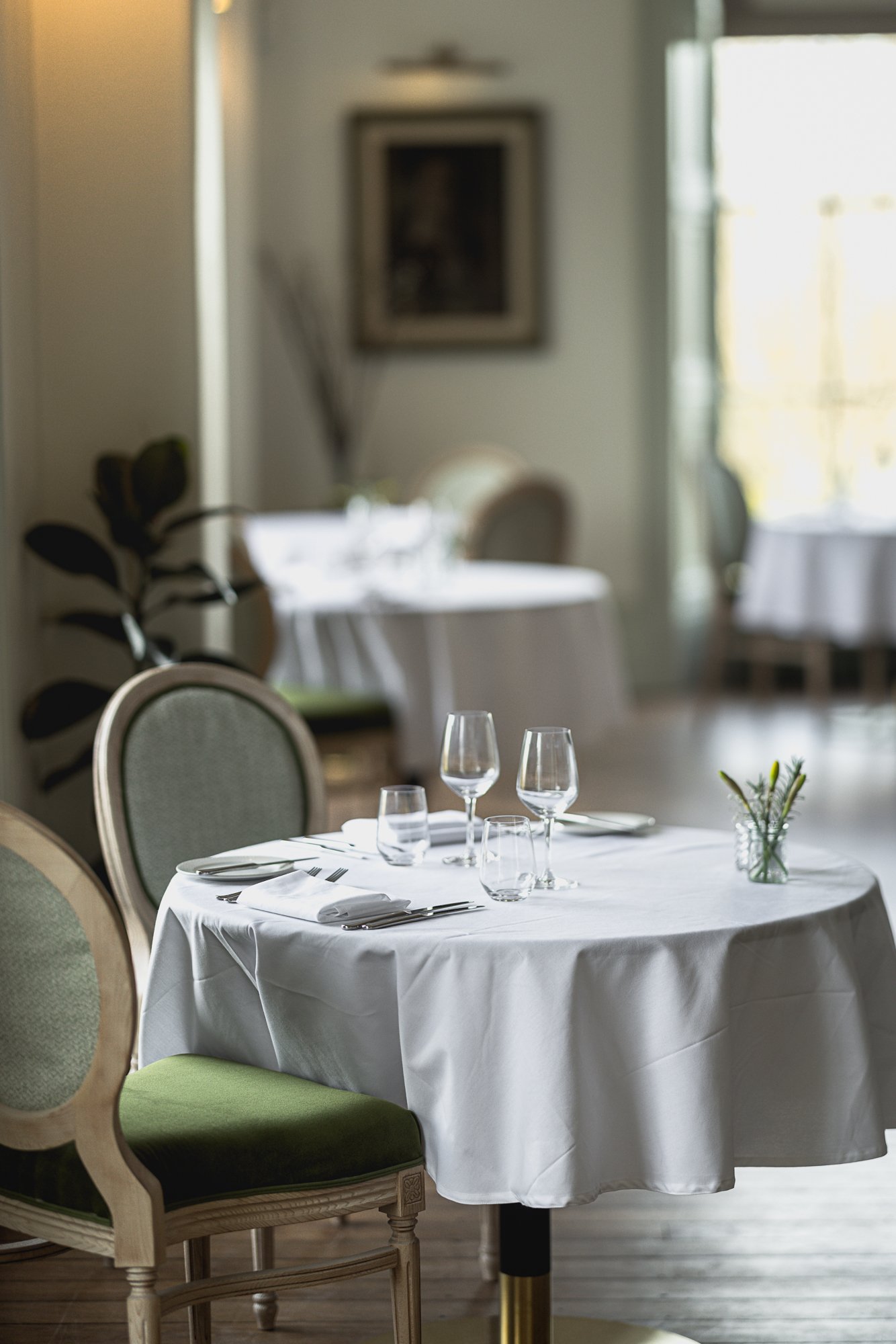 Fine-dining interior photograph at Mount Somerset in Taunton, showing a round table with white linen, glassware, cutlery and pale upholstered chairs against the dining room beyond.