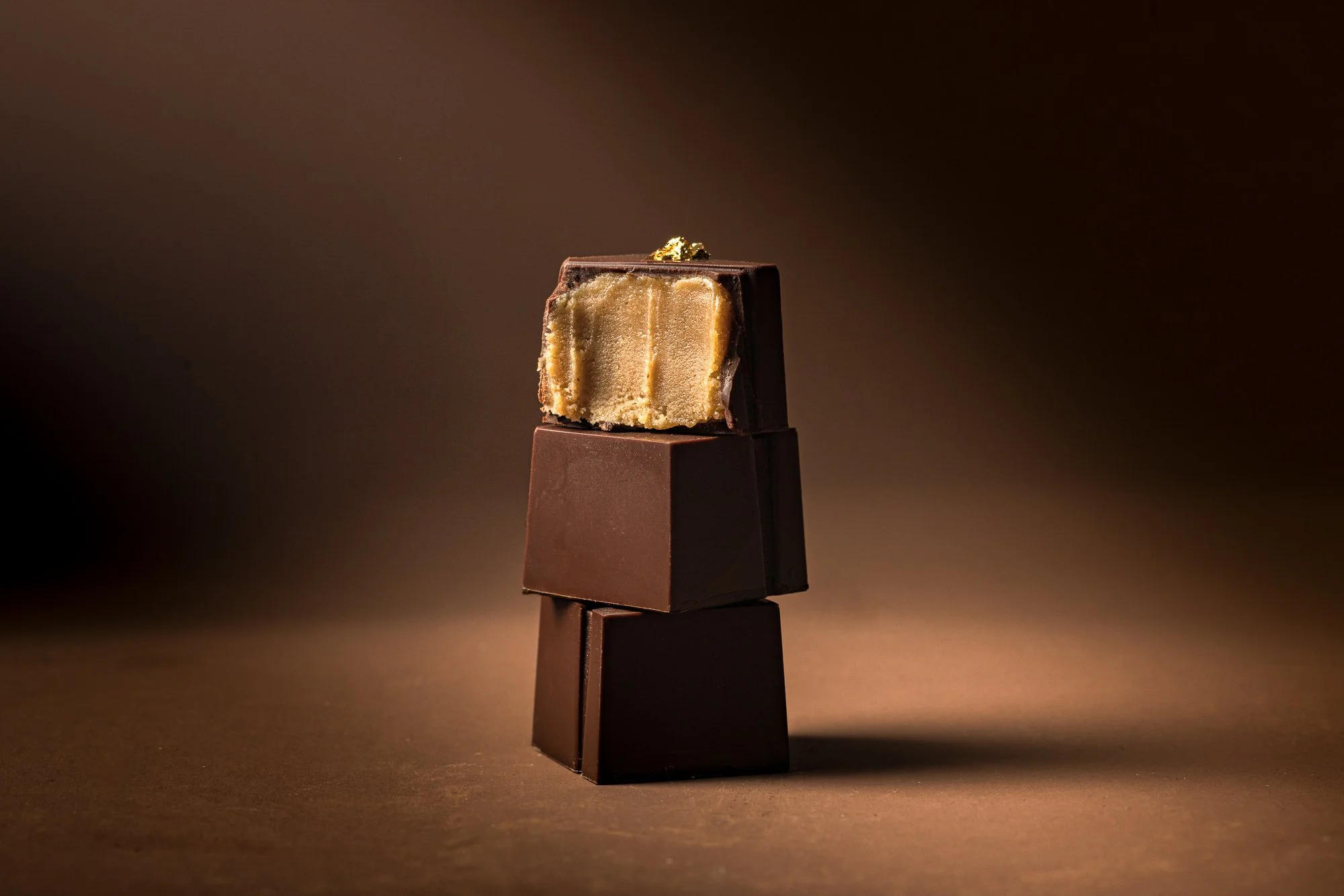 Studio chocolate photograph of a stacked dark chocolate confection topped with gold leaf against a softly graduated brown background, using controlled lighting to emphasise structure, luxury and surface detail.