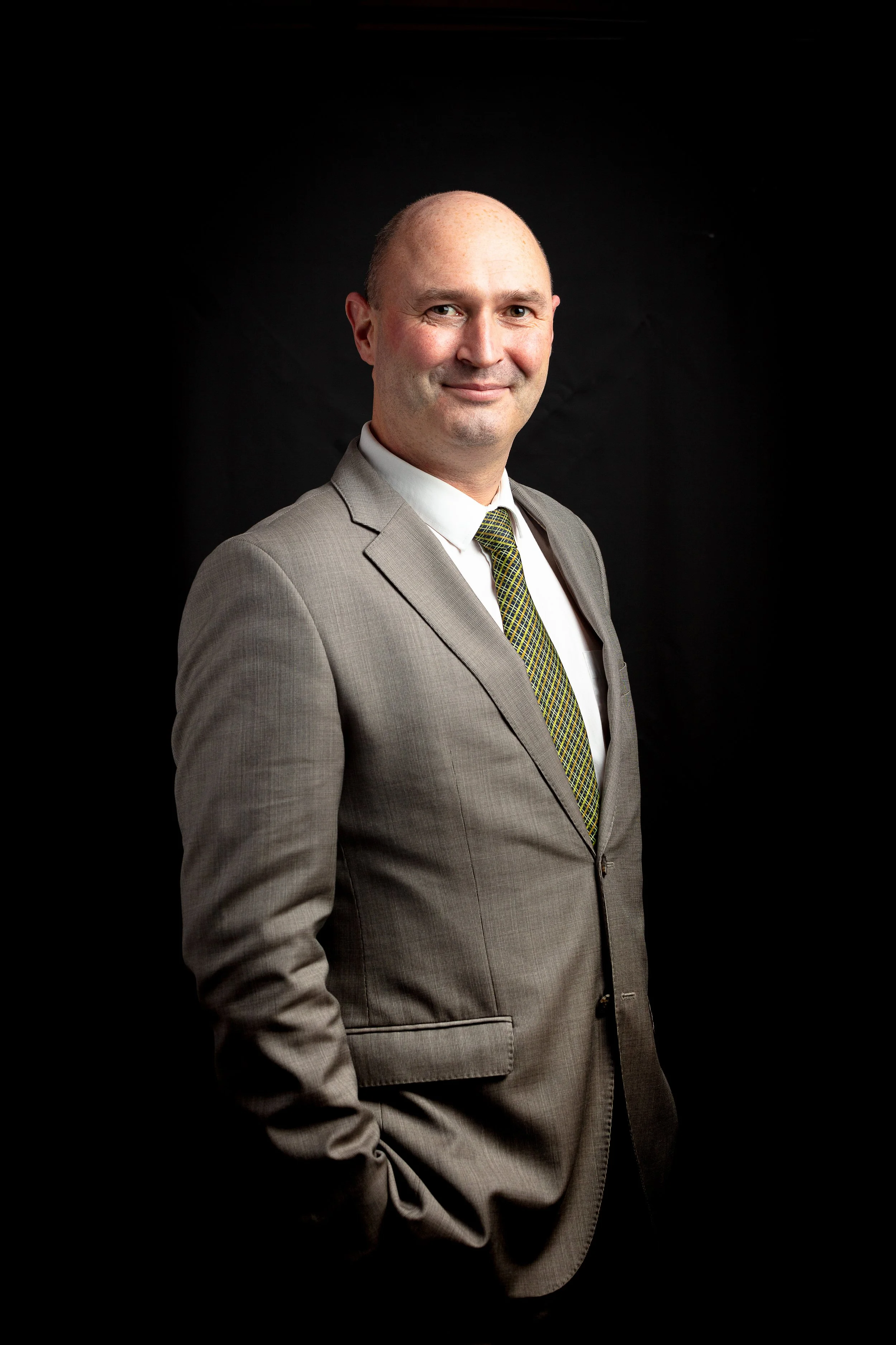 Formal corporate portrait of a businessman in a grey suit and tie against a black background, using controlled studio lighting and a relaxed standing pose for The Castle Hotel in Somerset