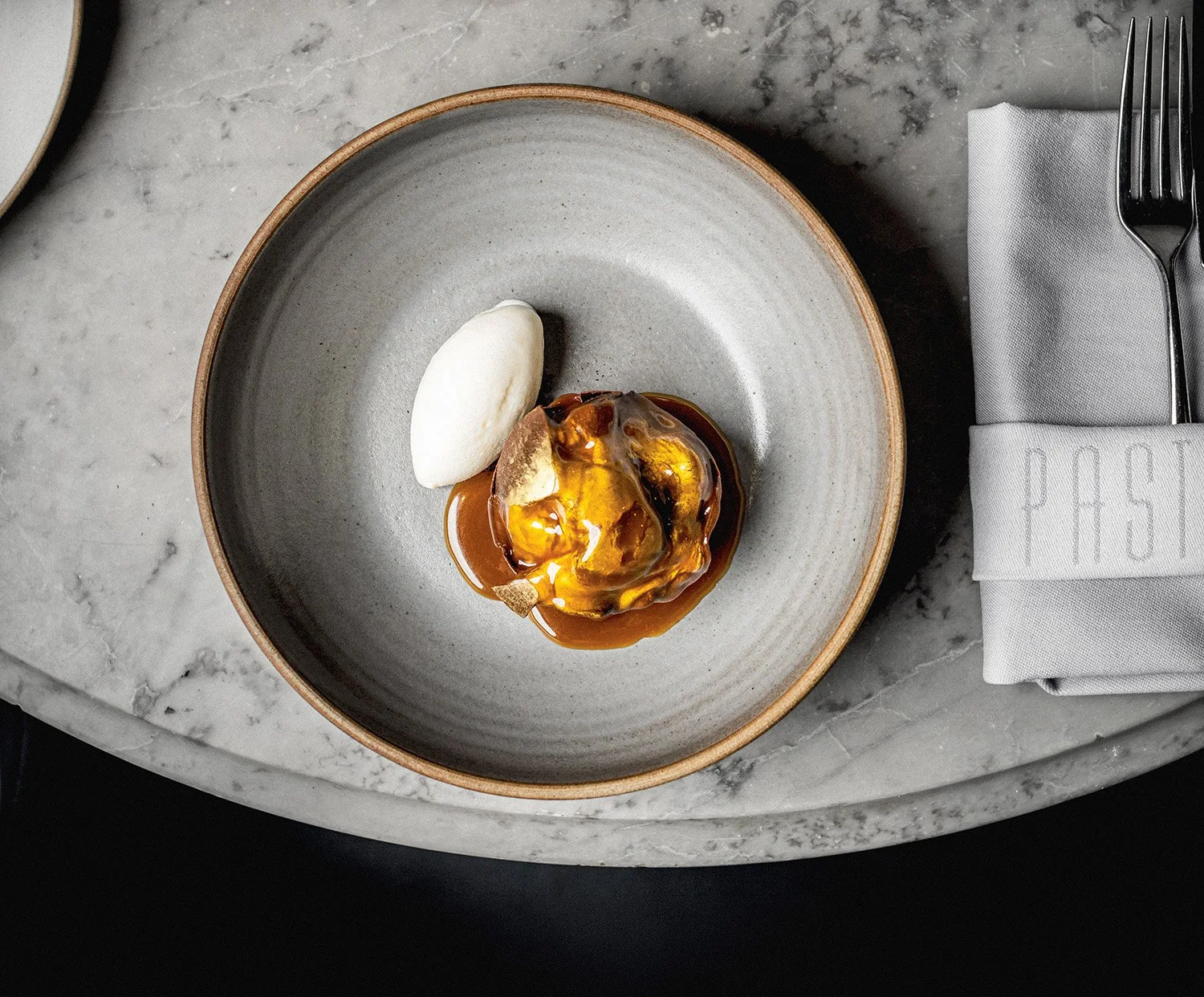 Overhead commercial restaurant photograph of a gold-dusted chocolate sphere melted open with hot caramel sauce, served with a white quenelle for Pasture Restaurants’ menus, websites and promotional use.