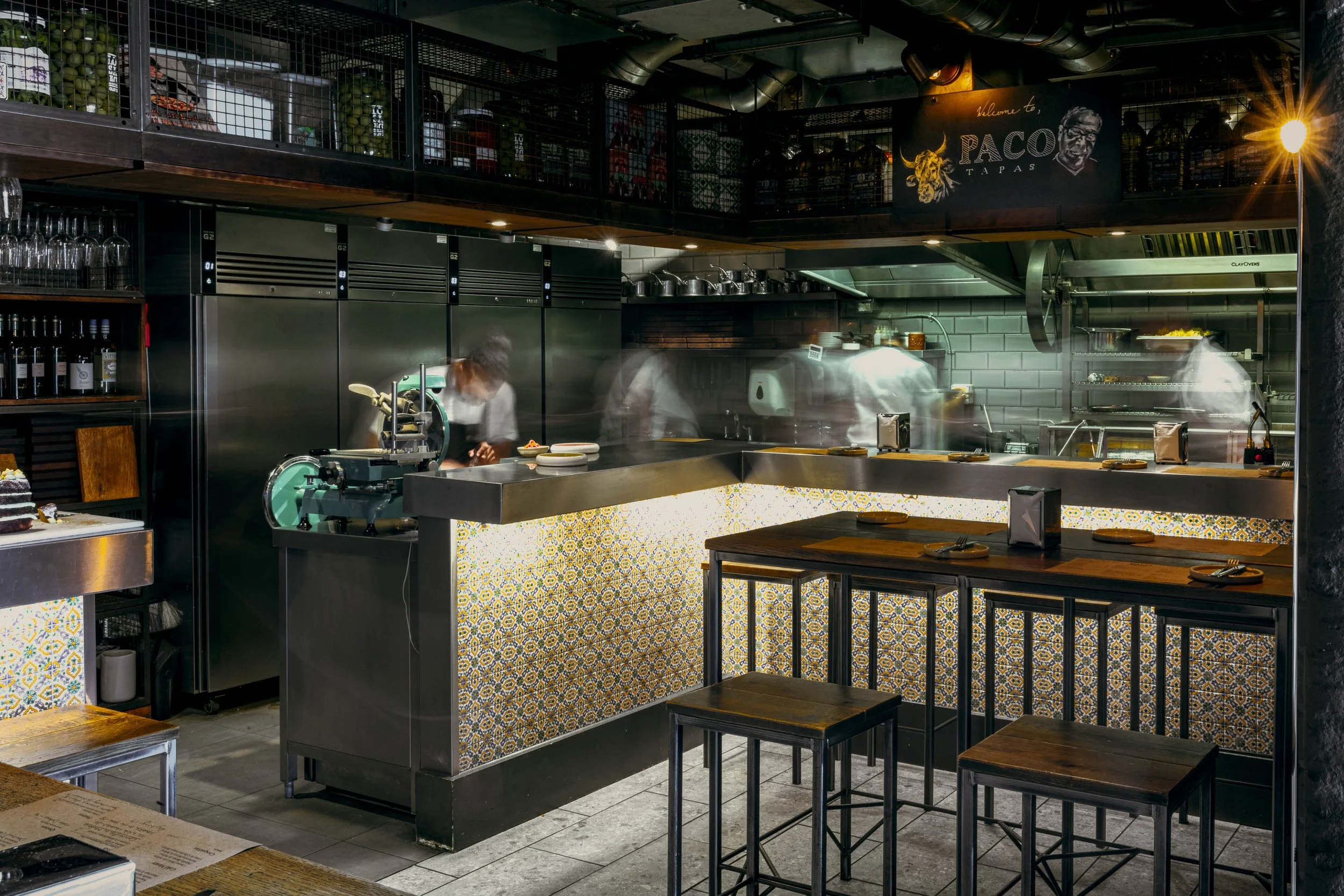 Editorial hospitality photograph of the open kitchen at Paco Tapas during service, with chefs rendered in movement behind the counter to communicate the pace, activity and working atmosphere of the Michelin-starred Bristol restaurant.