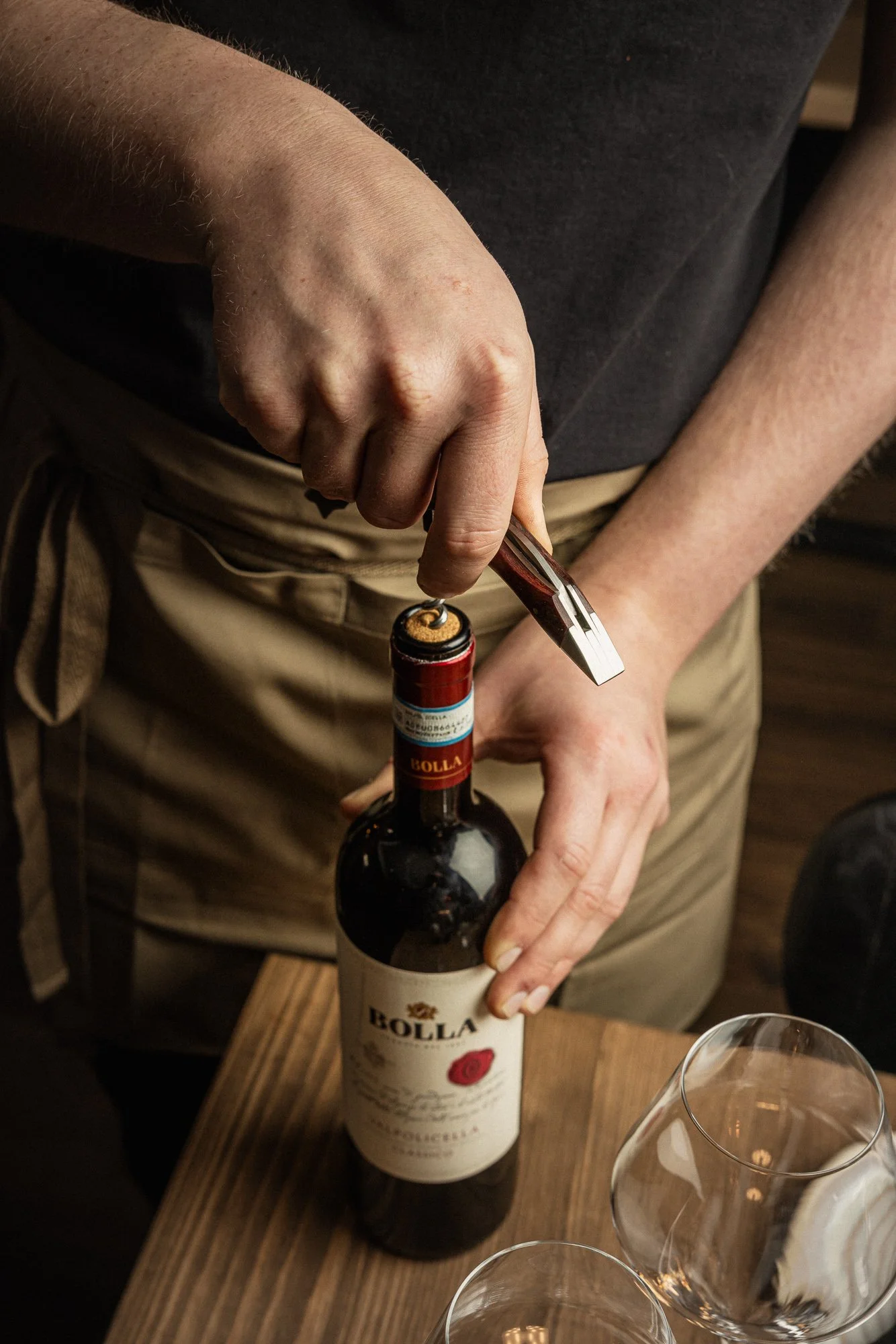 Close hospitality photograph of a server opening a bottle of red wine beside empty glasses, focusing on the hands, corkscrew and small practical gesture involved in restaurant wine service.