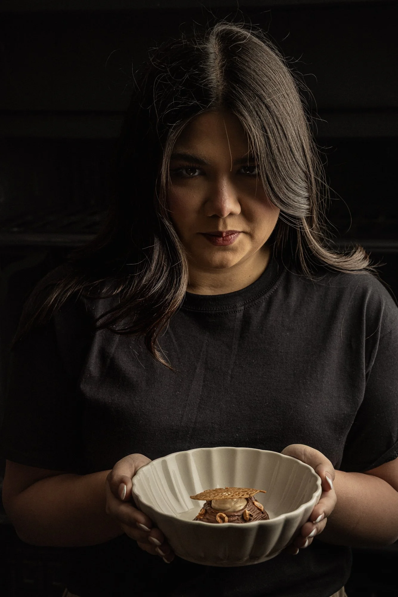 Atmospheric hospitality portrait of a woman holding a sculptural bowl containing a carefully plated dish, using restrained lighting and a direct gaze to connect the person with the food she is presenting.