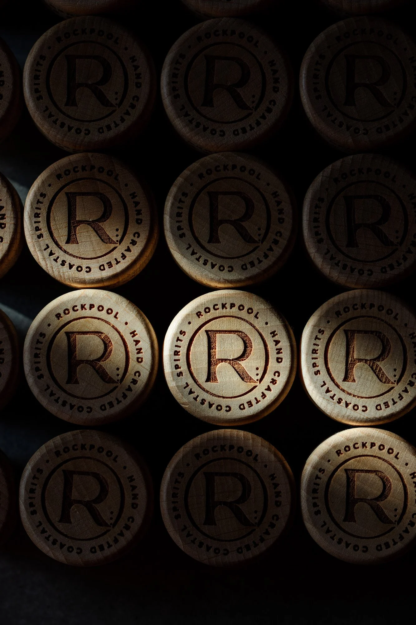 Graphic commercial photograph of repeating Rockpool Gin bottle closures arranged against a dark reflective surface, using controlled highlights and pattern to present the circular branding as a strong visual detail.
