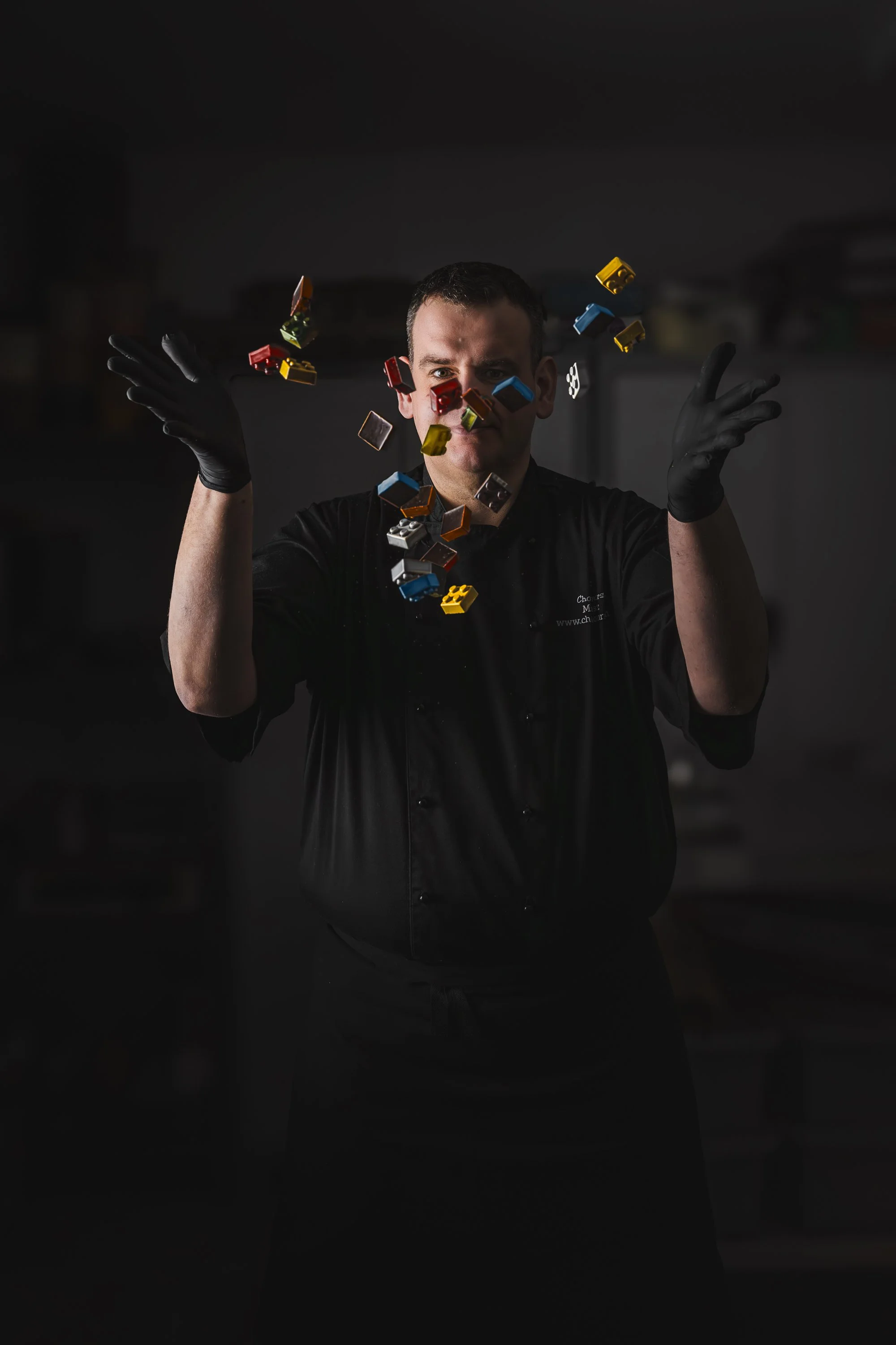 Creative commercial portrait of a chocolate maker dressed in black with colourful ingredients suspended around his face and hands, using controlled movement and a dark background to create a playful editorial image for Chocolours.