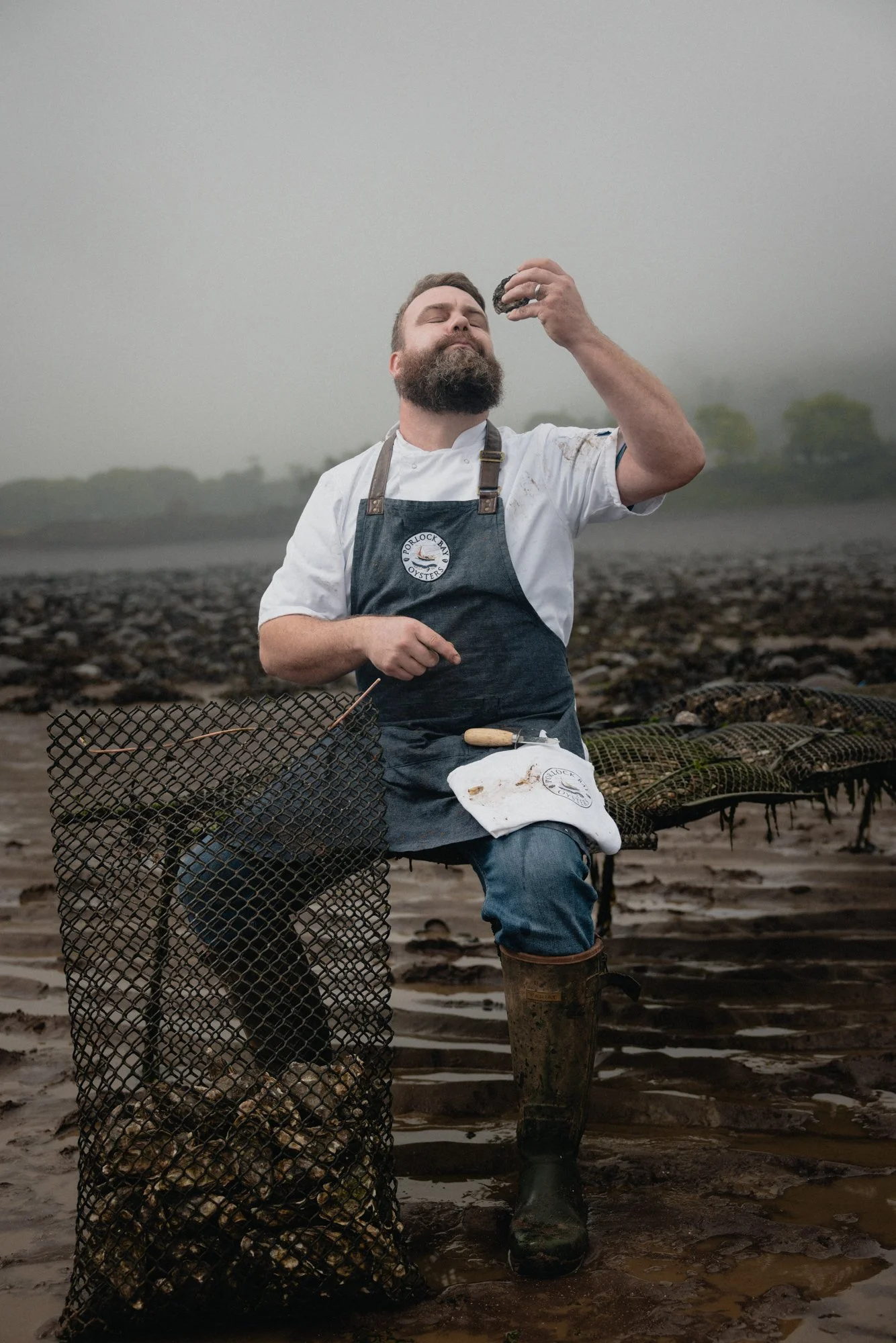 Documentary portrait of an oyster farmer carrying a heavy mesh basket of harvested shellfish through a working coastal site, recording the physical scale and practical conditions of oyster production for Porlock Bay Oyster in Somerset.