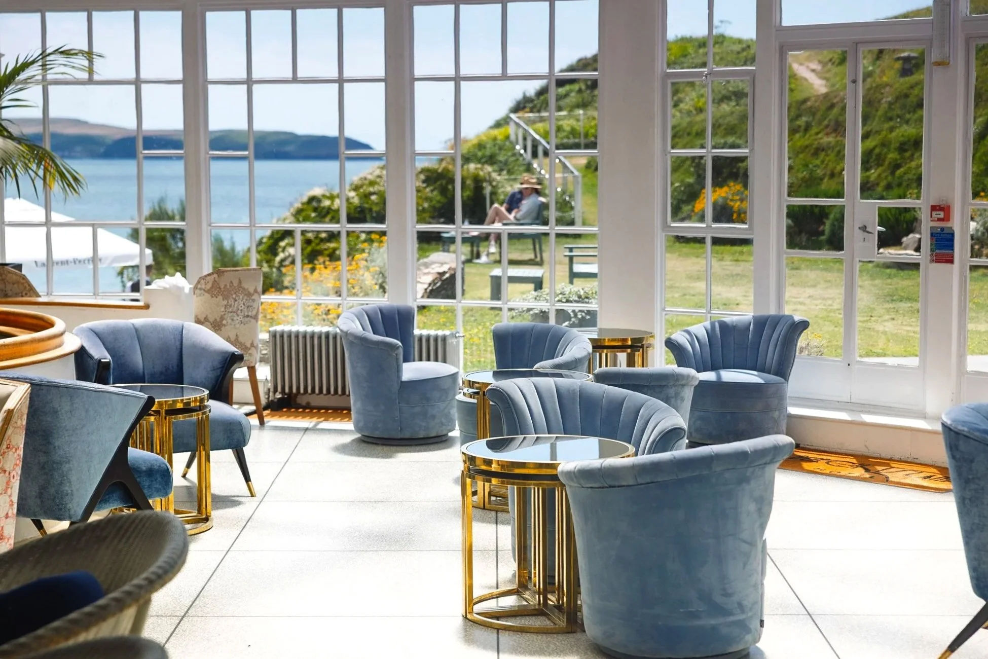 Bright commercial hotel interior at Burgh Island Hotel in Devon, showing pale blue armchairs, small gold tables and wide windows overlooking the sea and surrounding landscape.