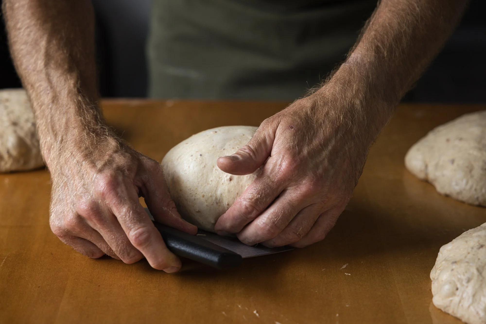 Close documentary food photograph of a baker’s hands shaping a round piece of dough with a scraper on a warm work surface, capturing the physical process and texture of bread production.