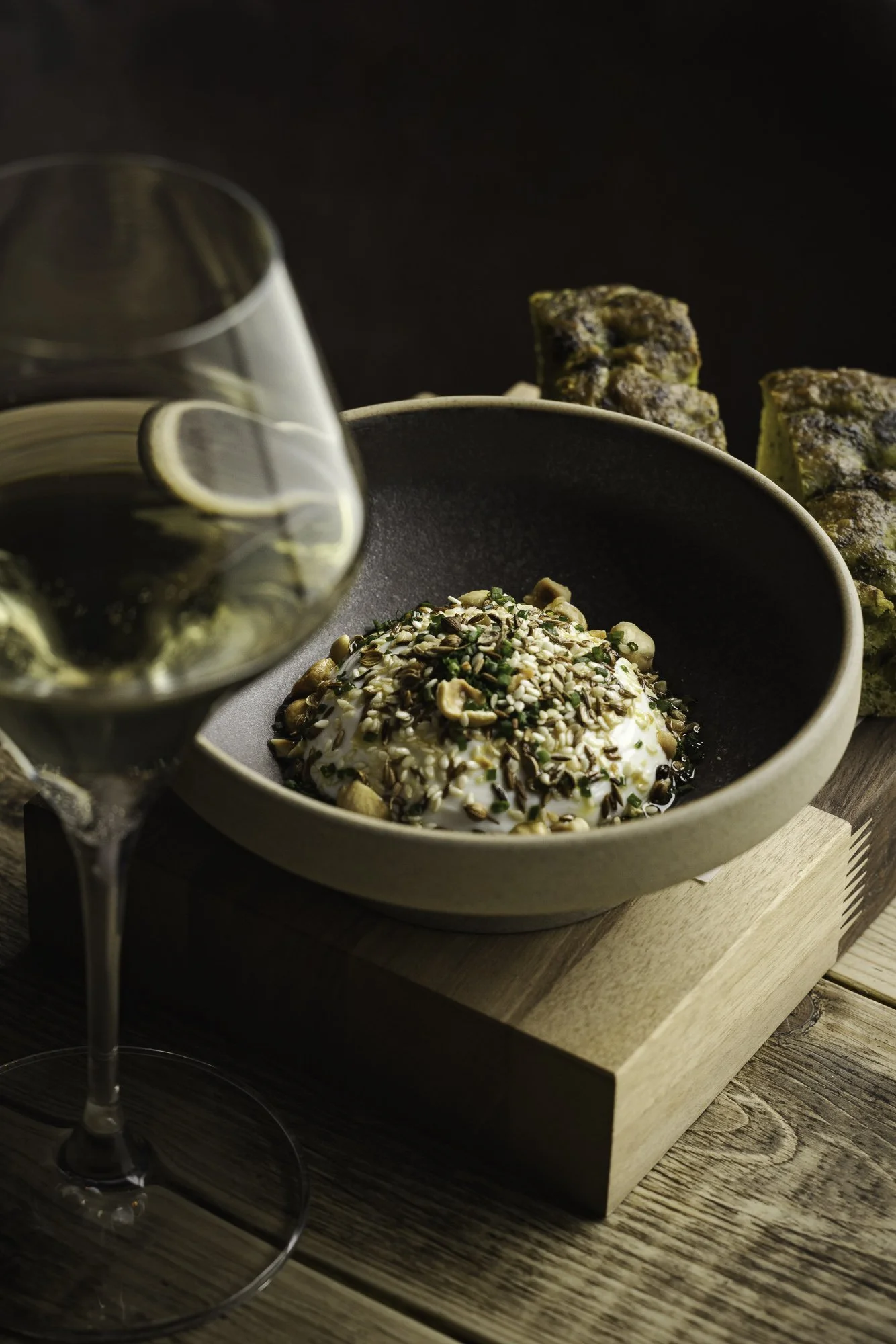 Restaurant food photograph of dish of humus   topped with mixed seeds and herbs in a dark ceramic bowl, framed by a softly focused wine glass and warm table light for restaurant or Kensington Restaurant  in North Devon