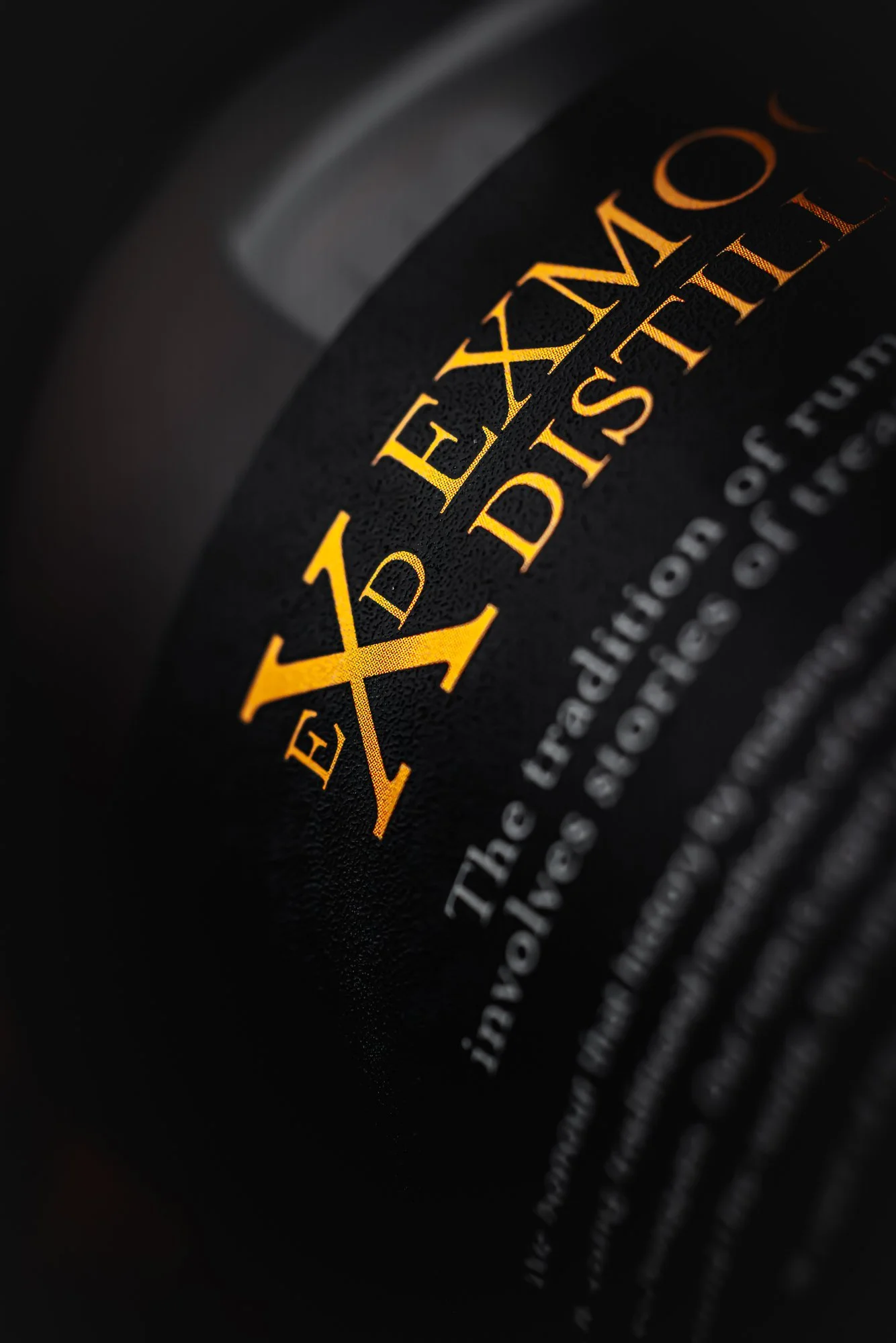 Macro commercial product photograph of an Exmoor Distillery bottle label with gold lettering against black glass, using shallow focus and directional light to emphasise the printing, typography and premium material finish.