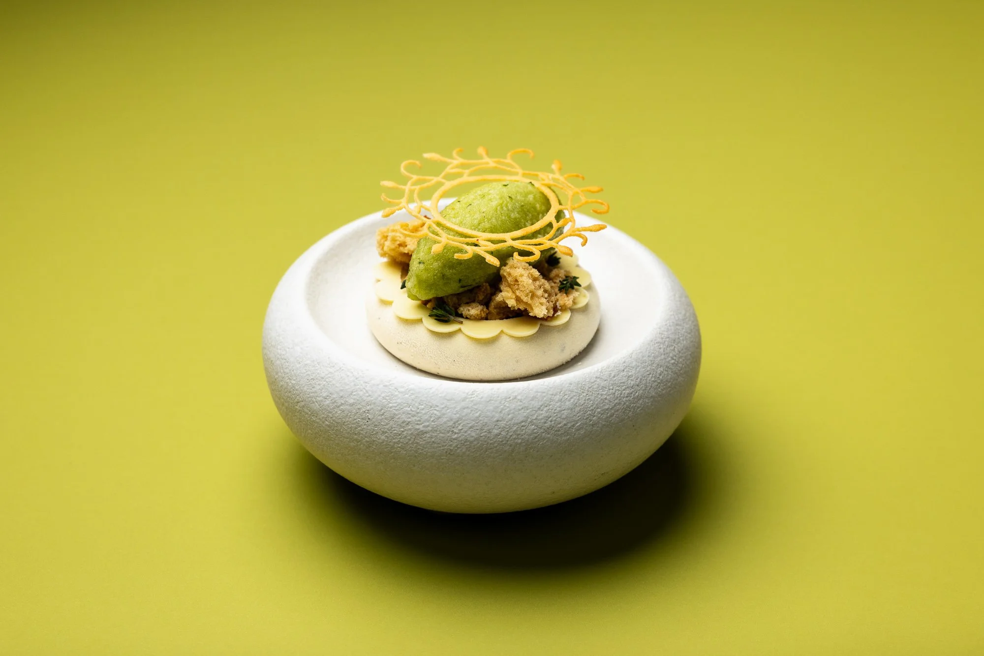 Commercial pastry photograph of an intricately plated green dessert with ice-cream, crisp decoration and layered components on a white plate, using a vivid lime background for Pastry Academy UK by David Vidal.