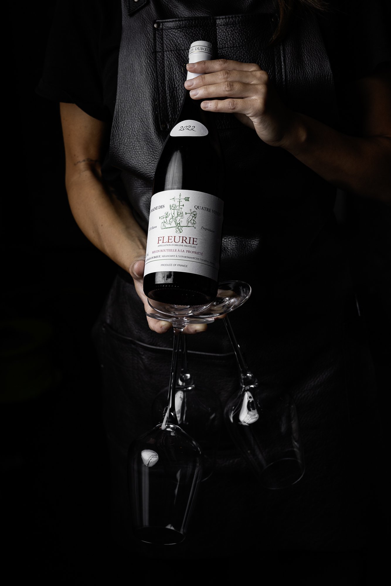 Commercial wine photograph showing a server pouring Fleurie from the bottle into a large stemmed glass, with the hands, label and falling red wine isolated against a dark background for Castle Hotel Taunton.