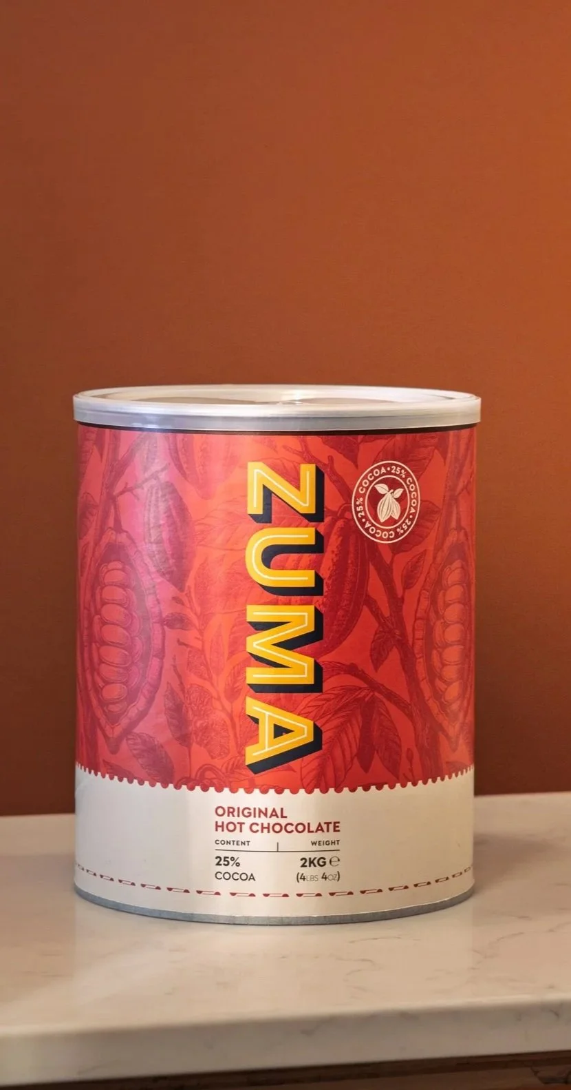 Commercial product photograph of a red Zuma Original Hot Chocolate tin presented upright against a warm terracotta background, clearly showing the packaging, branding and retail format.