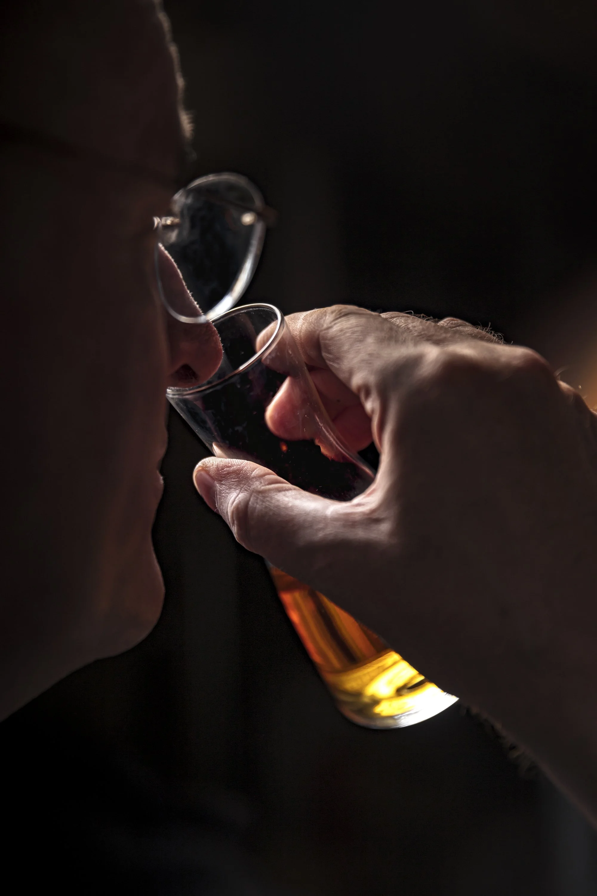 Close environmental portrait of a drinks producer holding a small glass of amber spirit towards the light for inspection, focusing on the eye, hand and liquid to communicate the care involved in assessing the finished product.