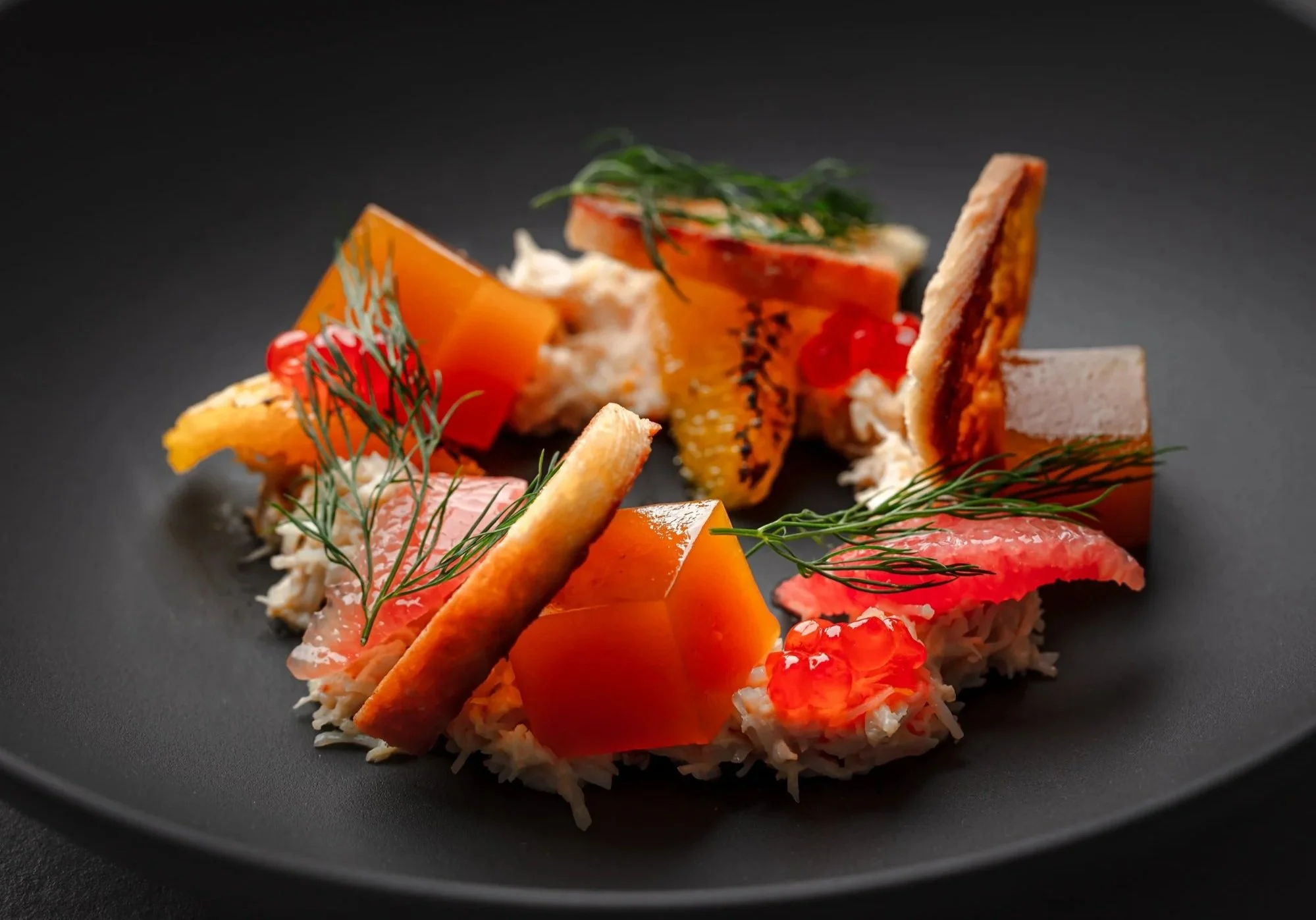 Restaurant food photograph of crab arranged with vivid orange and red ingredients, herbs and crisp elements against a dark background, using precise lighting to emphasise colour, texture and presentation. Shot for Dine Indulge in Bristol
