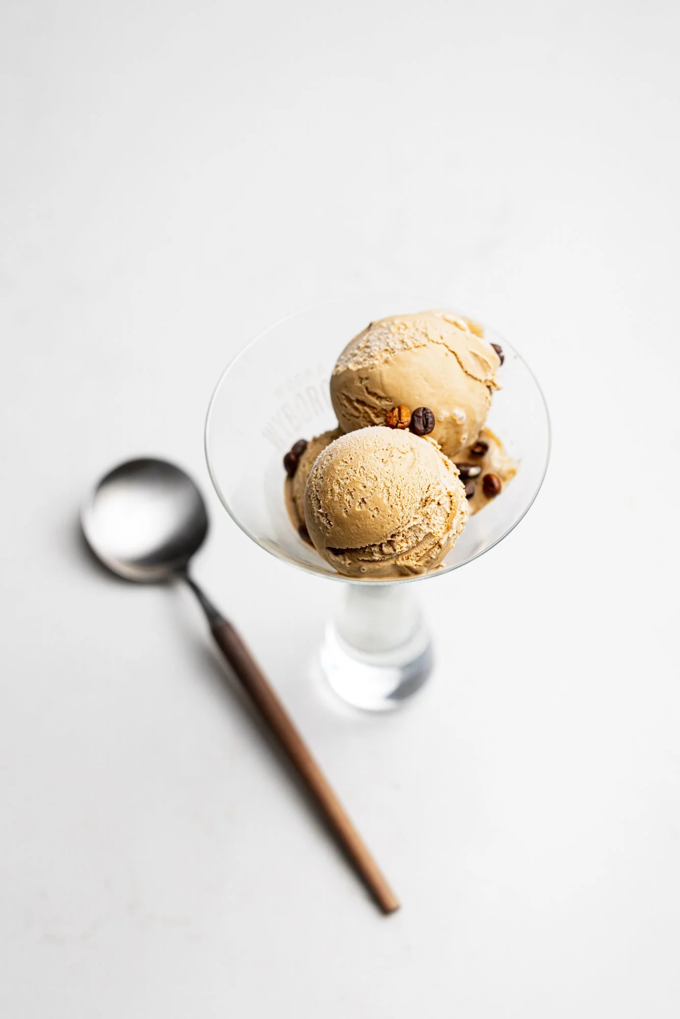 Clean commercial food photograph of three scoops of Caramel ice-cream flavour served in a clear dessert glass with a spoon, using a bright white background and restrained composition for Berties Ice-cream.