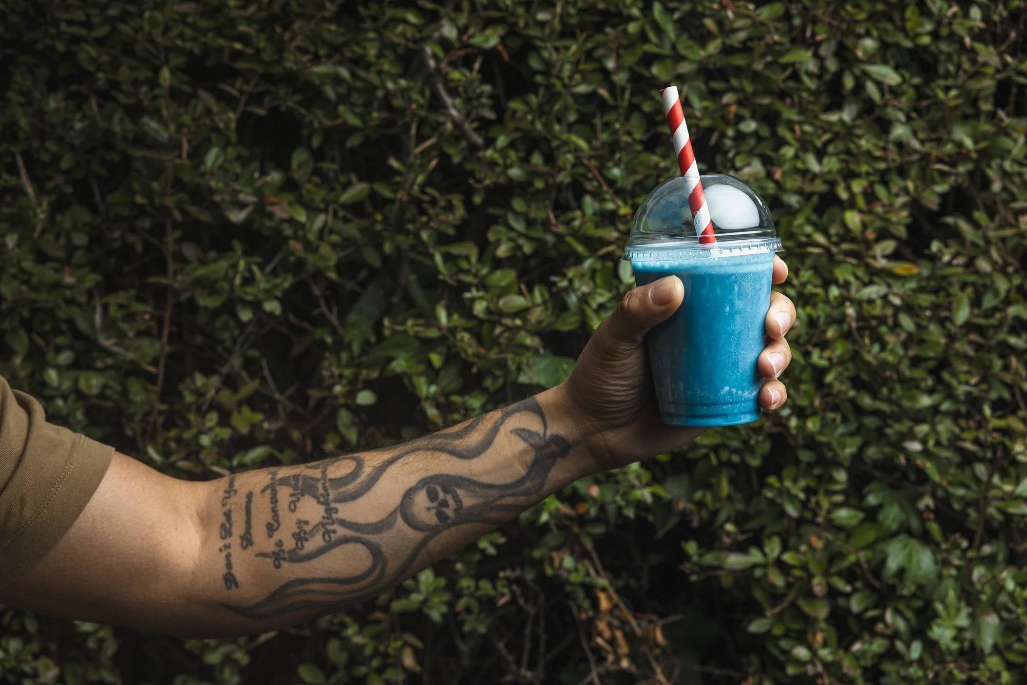 Lifestyle drink photograph of a tattooed arm holding a bright blue frozen drink with a striped straw outdoors, using dense green foliage as a contrasting background for Coffee Number 1 summer campaign.