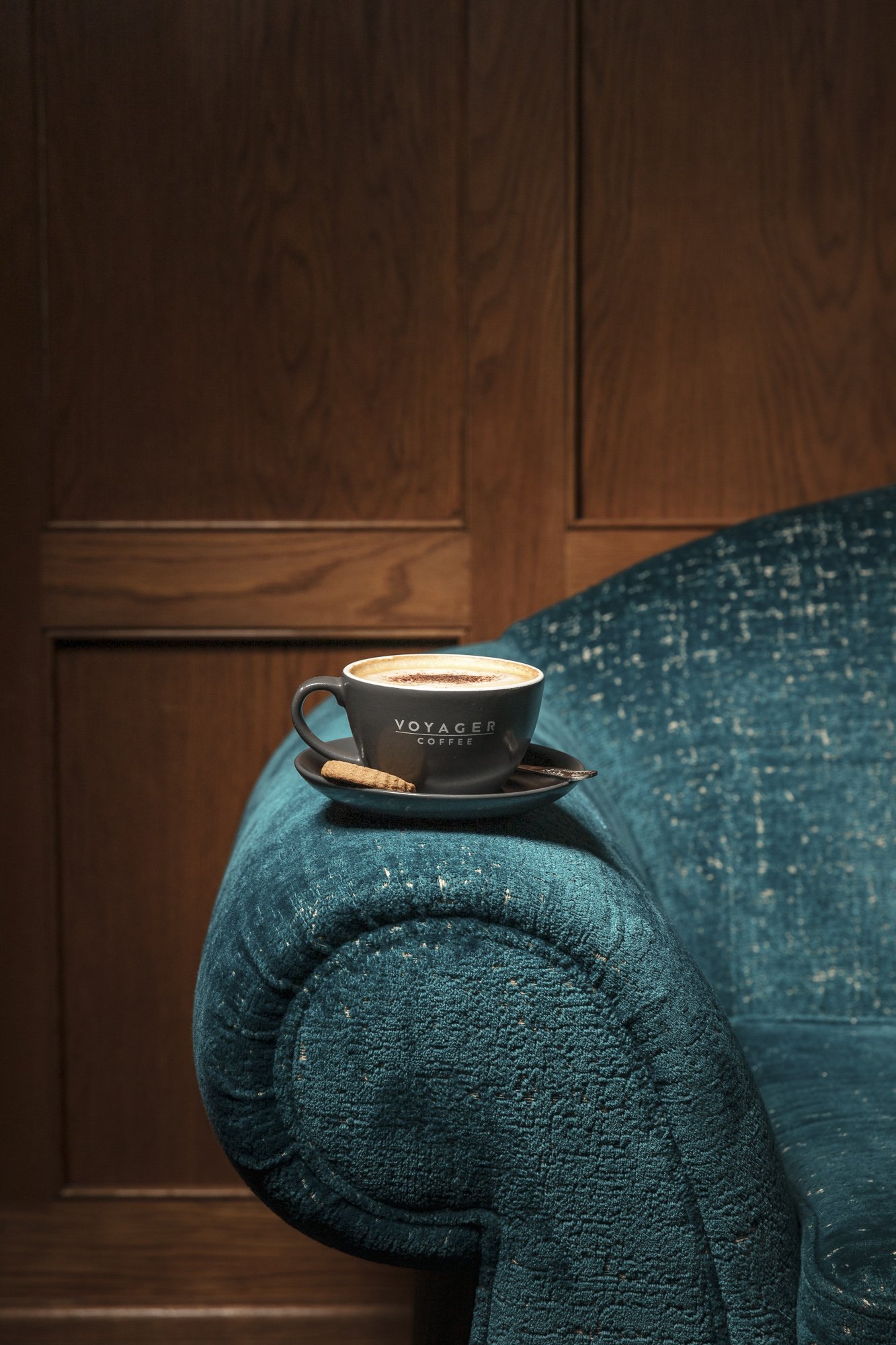 Interior-led commercial coffee photograph of a Voyager Coffee cup resting on the curved arm of a teal upholstered chair, using warm wood panelling and rich fabric texture to place the drink within a relaxed hospitality setting.