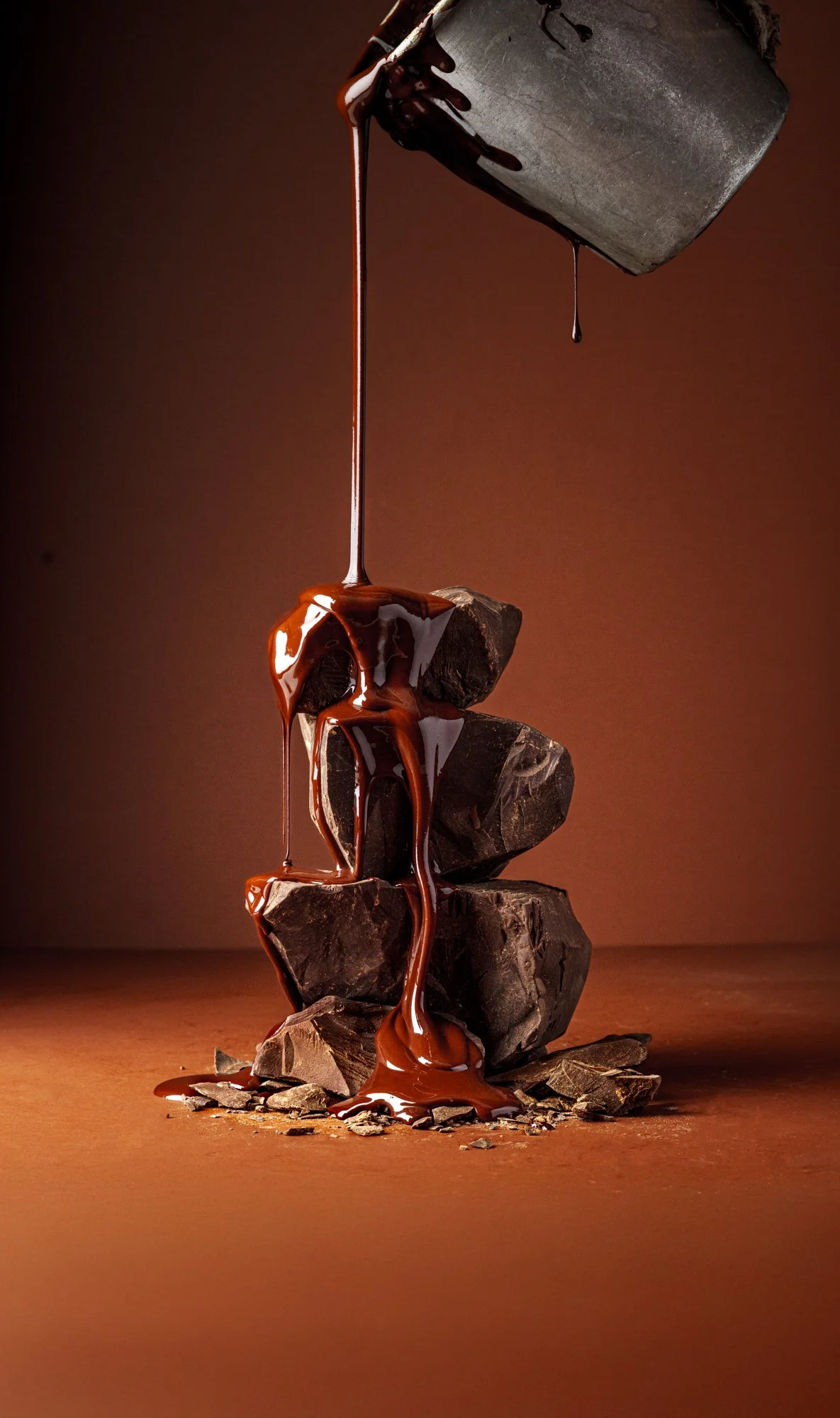 Commercial food photograph of glossy chocolate pouring over a stack of irregular chocolate pieces against a warm brown background, freezing the liquid movement and contrasting textures in mid-flow.