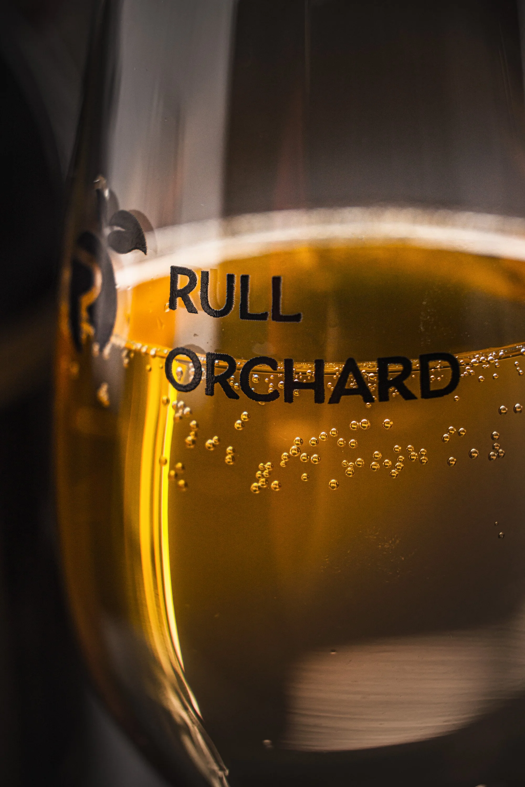 Close commercial photograph of golden Rull Orchard cider in a branded glass, using warm backlighting to reveal rising bubbles, colour and printed lettering associated with the Somerset cider producer.